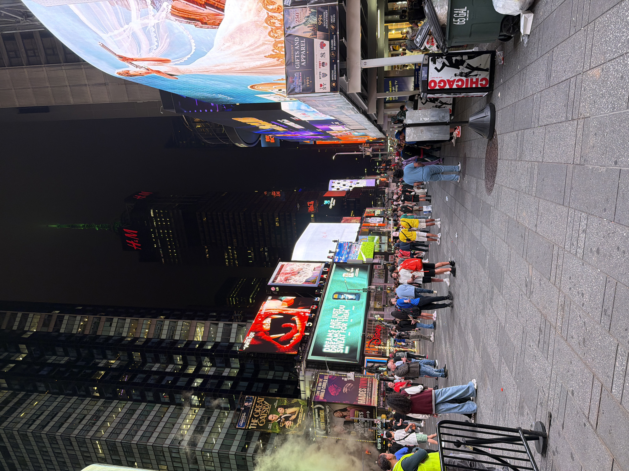 This is a nighttime, eye-level, wide shot of Times Square in New York City, USA. The scene is bustling with a crowd of people walking on the paved ground. Buildings with illuminated windows tower over the square, adorned with large, bright digital billboards advertising various products and shows. One billboard prominently features a close-up of a red, fanged mouth, another shows a vintage poster for "The Great Gatsby" musical, and a third displays a woman in a white dress with a veil. The iconic H&M building is visible in the background, topped with a green spire. The overall atmosphere is one of urban vibrancy and commercial activity, illuminated by the glow of neon signs and screens. Several trash receptacles are present, one of which is a green 65-gallon bin with "CHICAGO" written vertically on a nearby advertising panel.