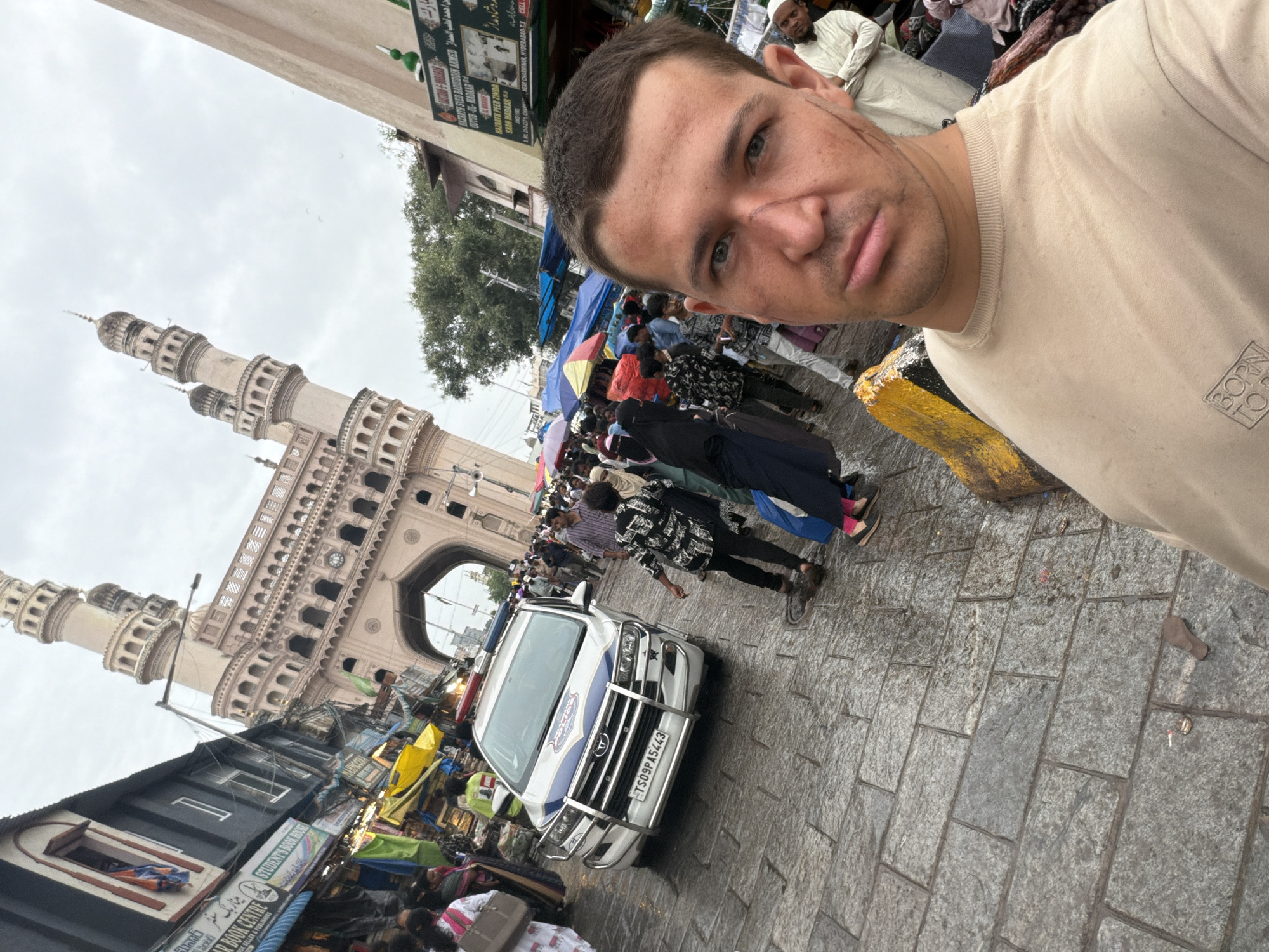 The image is a selfie taken in front of the Charminar in Hyderabad, India. The Charminar, a historical monument and mosque, is visible in the background with its distinct four minarets and arched gateway. In the foreground, a man is taking the selfie. He has visible scars on his face. A white police vehicle with blue and red lights on top is parked on the cobblestone street. The street is crowded with people, some are dressed in traditional Indian attire. There are market stalls with colorful awnings lining the street, indicating a busy commercial area. The ground appears wet, suggesting it might have rained recently.