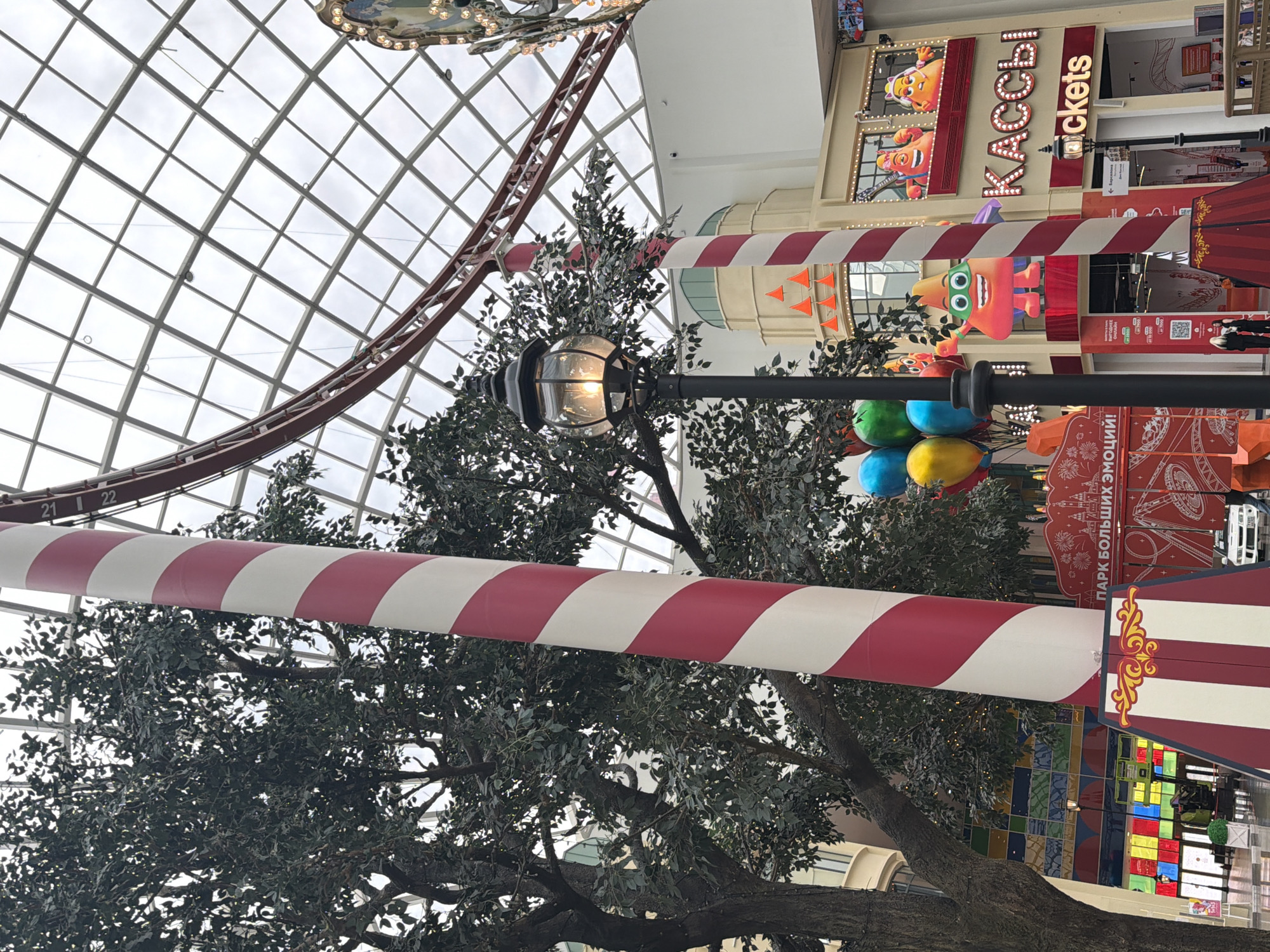 This is a medium-wide, eye-level shot of an indoor amusement park, likely a shopping mall, with a transparent, grid-like roof. In the foreground, a large, red and white striped candy cane-like pole dominates the left side of the frame. To its right, a street lamp with a glowing bulb illuminates the scene. Behind the pole, a large, artificial tree with dark green foliage is visible. Further in the background, a red roller coaster track curves across the ceiling.
A cluster of colorful balloons, red, yellow, and blue, floats to the left of the lamp post. A brightly lit storefront with the Russian word "КАССЫ" (KASSY), meaning "TICKETS," is visible on the right, with animated figures above. Another similar striped pole is present behind the ticket booth. In the lower left, a section of colorful tiled wall and a display of merchandise are visible. Two women in dark clothing stand near the bottom center, conversing. In the far right, a portion of a carousel is visible. The overall atmosphere appears festive and commercial, with bright lights and playful decorations.