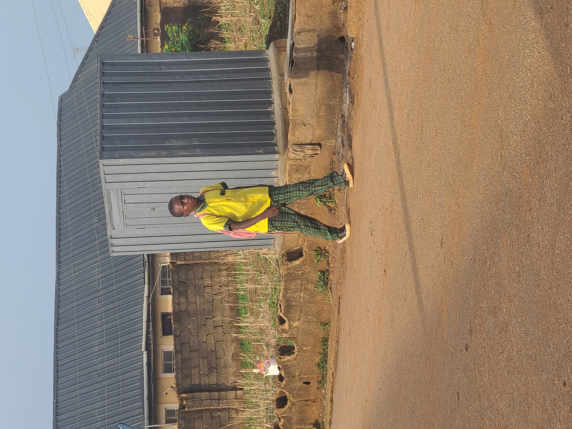 A person wearing a yellow shirt and plaid pants stands on a paved surface, possibly a street, in what appears to be a residential or urban area of Jos, Nigeria. The person has a red bag slung over their shoulder. Behind them is a structure with a gray metal roof and walls. There's also a brick wall and some vegetation. The paved surface takes up a large portion of the foreground. Overhead power lines are visible against the blue sky.