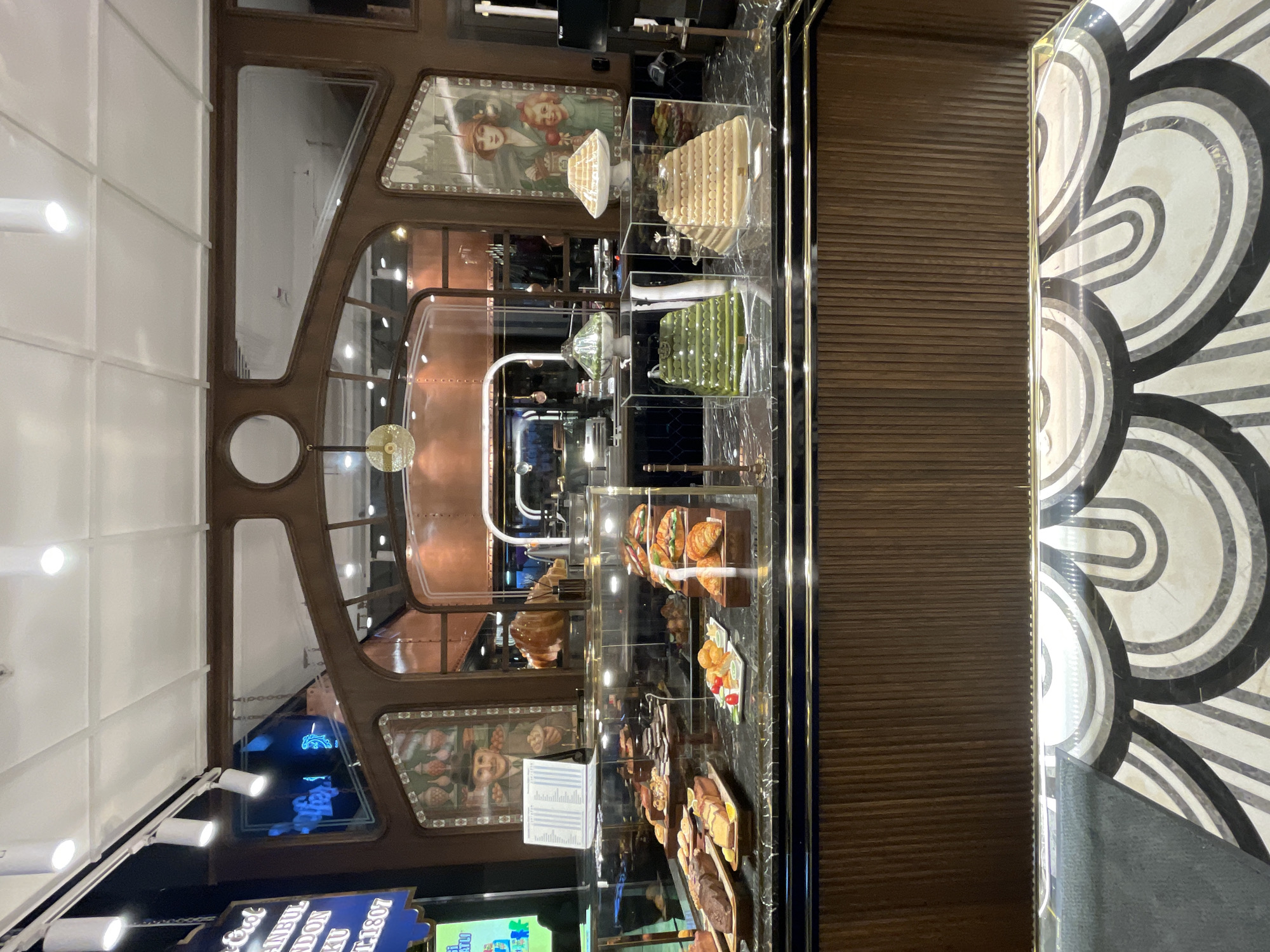 A confectionery display case is shown in a retail establishment, likely in Kadıköy, Turkey, with various pastries and desserts arranged on marble countertops. The display case is made of glass and brass, with wooden paneling on the exterior. Above the display case, there is a decorative wooden frame with mirrored sections and a hanging lamp. Behind the display, a large pastry, possibly a croissant, is visible. Two tiered desserts, one green and one golden brown, are presented in clear acrylic boxes. The background shows a retail setting with lighting and reflective surfaces. The floor in front of the counter features a black and white geometric pattern. The overall ambiance suggests a high-end patisserie or bakery.
