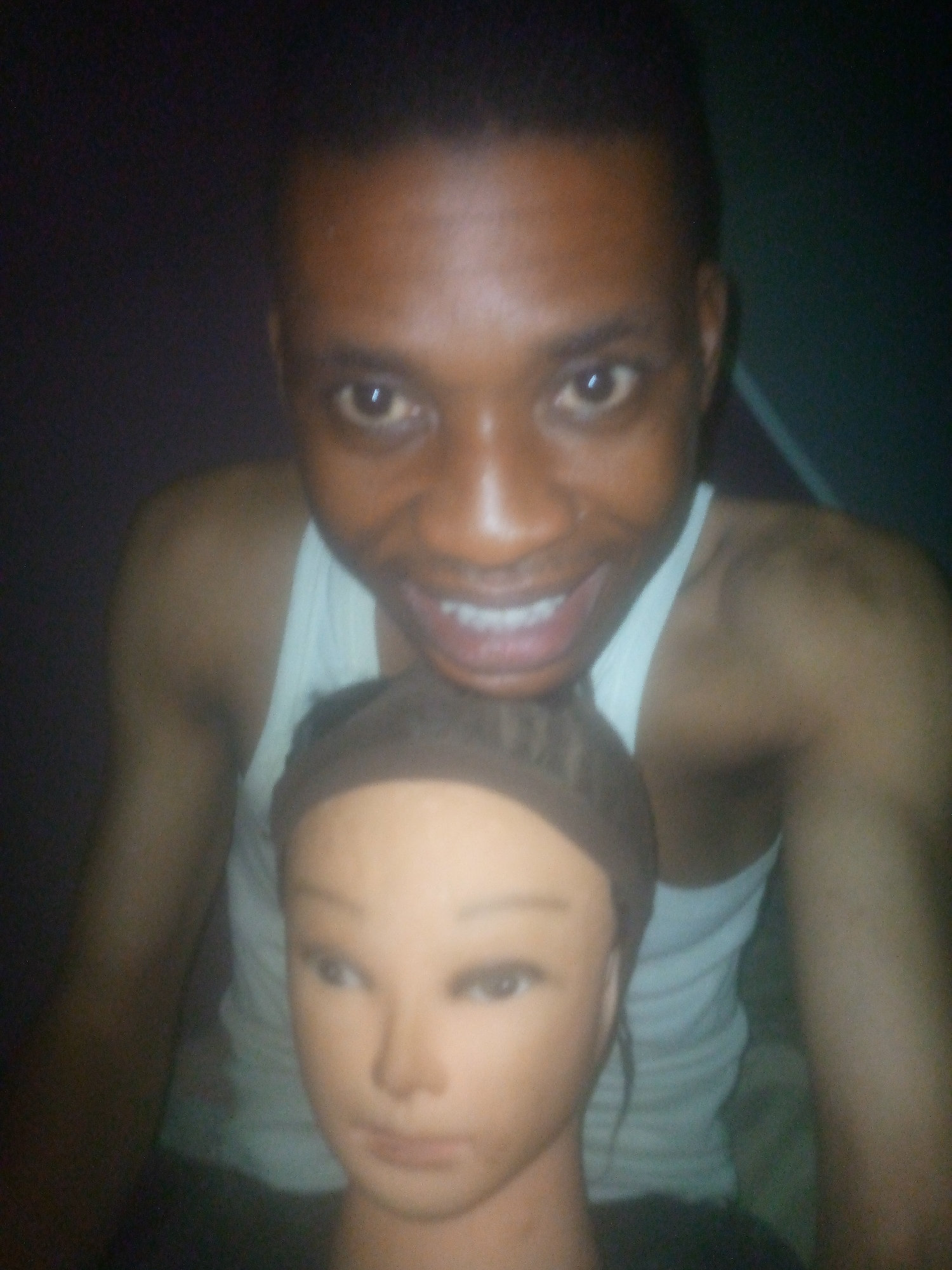A close-up shot shows a man wearing a white tank top and smiling broadly. He is standing behind a mannequin head. The location is unknown. The environment appears dimly lit.