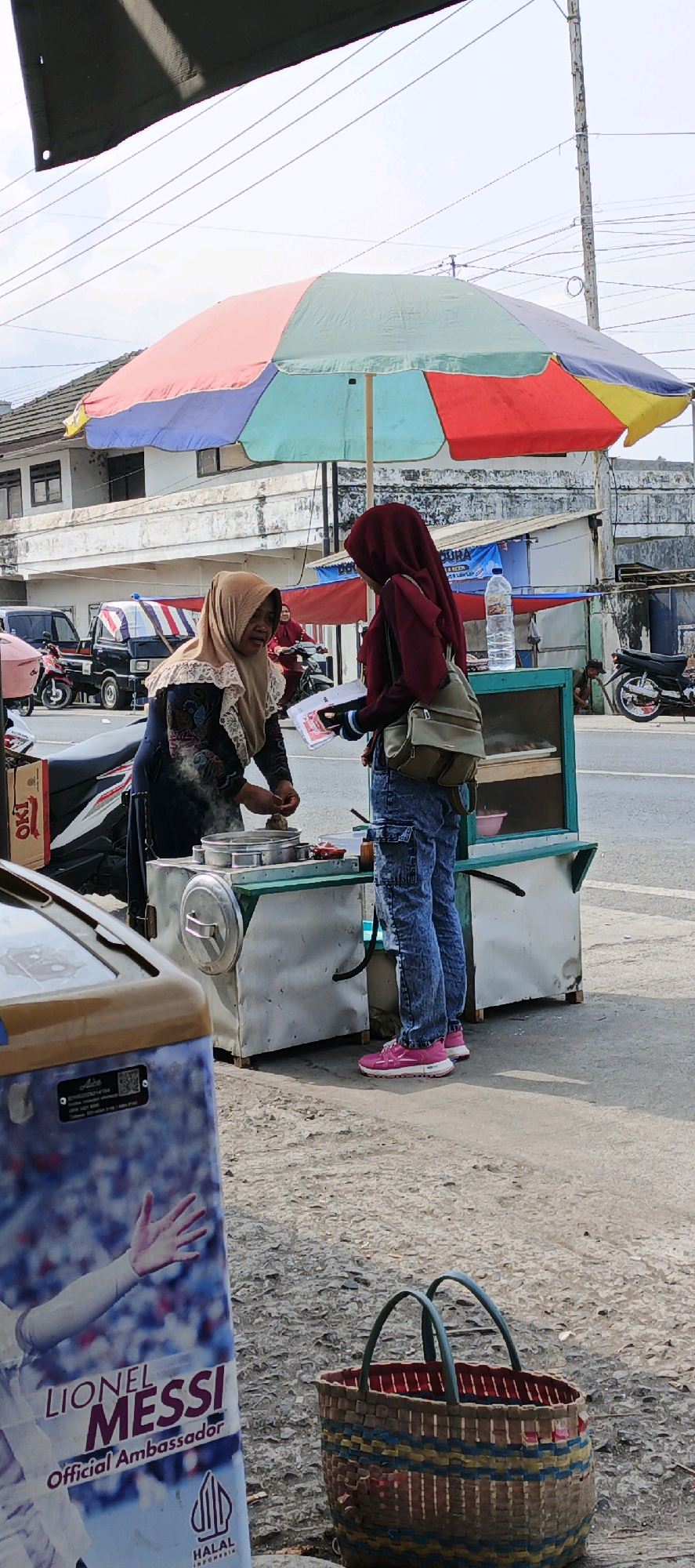 In Kasiyan, Indonesia, an outdoor scene unfolds under a vibrant, multi-colored umbrella. Two women, both wearing hijabs, are engaged in a transaction. One woman, in a tan hijab and patterned blue dress, stands behind a food cart, actively preparing food. The other woman, in a maroon hijab and distressed blue jeans, faces her, holding what appears to be money and a pamphlet or menu. A green backpack is slung over her shoulder. The food cart, made of metal with a green trim, displays some food items behind glass. To the left, a portion of a refrigerated display is visible, featuring an advertisement with a hand and the text "LIONEL MESSI Official Ambassador" and a "HALAL INDONESIA" logo. In the background, parked vehicles, including a black pickup truck and motorcycles, suggest a roadside setting. A utility pole with numerous wires is also prominent in the background. The ground is paved with asphalt and gravel. A woven basket with red and blue accents rests on the ground in the foreground.