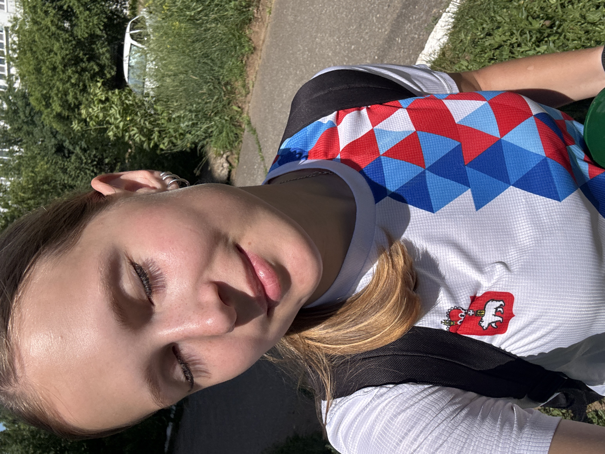 The image shows a young woman with blonde hair pulled back in a ponytail, wearing a white t-shirt with a colorful geometric pattern on the right side and an emblem of a white bear on a red shield on the left side. She is wearing a black backpack. Her eyes are closed and she is smiling. She has a silver hoop earring in her left ear. The background consists of green trees, tall grass, a paved path, and a white car. This scene is located in the city of Chaikovsky, Russia.