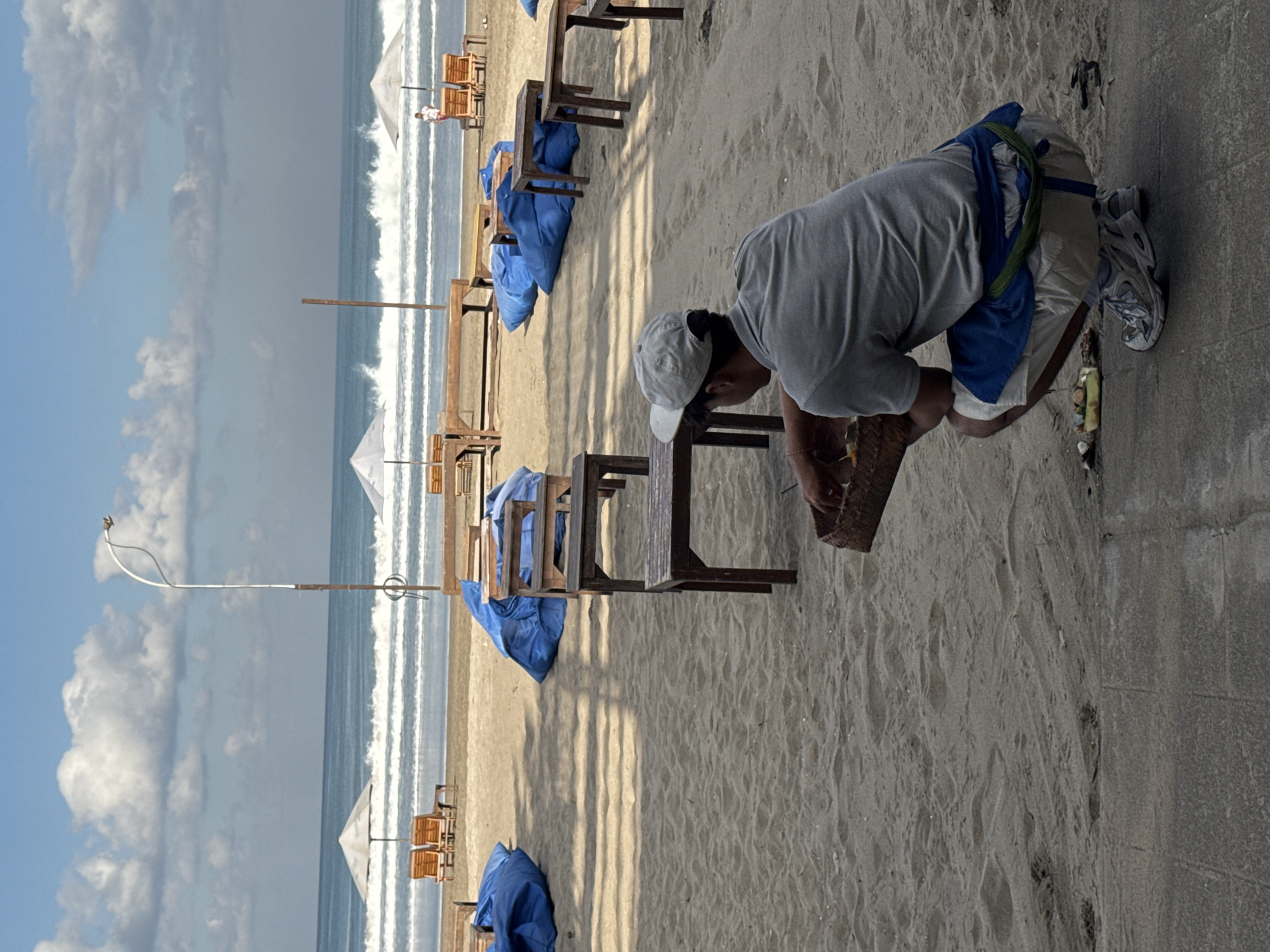 This detailed image captures a beach scene in Kuta, Bali, Indonesia, under a partly cloudy sky. The ocean, characterized by breaking white waves, occupies the background horizon. The sandy beach in the mid-ground features several white beach umbrellas, each accompanied by two wooden sun loungers. Additional wooden tables and large blue, possibly cushioned, seating elements are scattered across the sand. A tall, curved metal pole, appearing to be a light standard, is positioned on the right side of the beach area.
In the foreground, a paved walkway separates the viewer's immediate space from the sandy beach. A man is observed squatting on this paved surface in the bottom right quadrant, facing towards the beach with his back partially to the camera. He wears a light grey short-sleeved shirt, light-colored shorts, a blue fabric tied around his waist, and a green cord secured over the blue fabric. A light grey baseball cap is worn backward on his head, and he is wearing white and blue athletic shoes. He holds a woven rectangular basket, containing small items, over a small cluster of debris on the pavement. A yellow rectangular container with additional items is positioned near his right foot on the ground. The scene is illuminated by natural daylight, casting shadows on the sand.