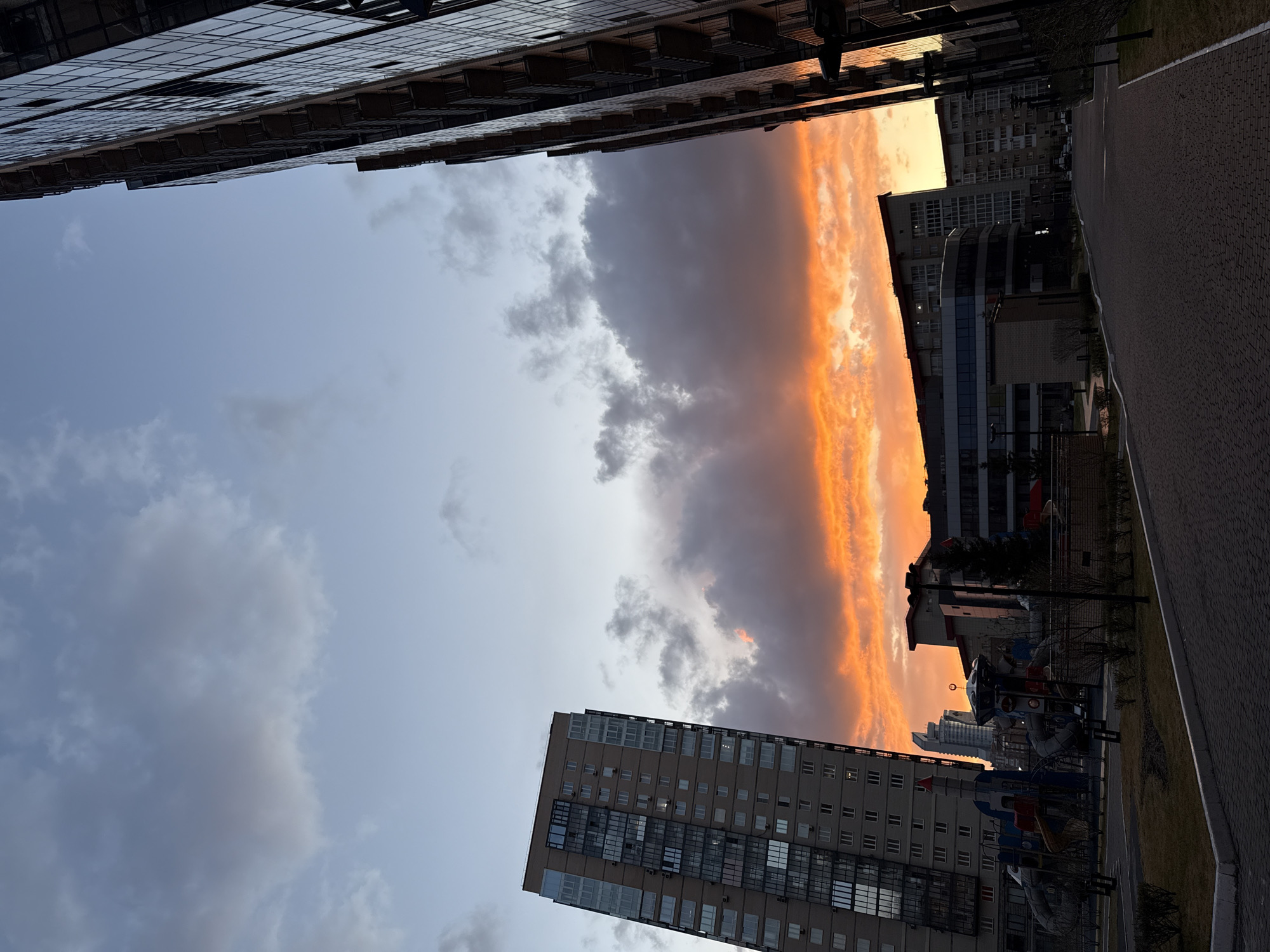 The image shows an upward-looking perspective of a street in Krasnoyarsk, Russia, at sunset. Tall residential buildings are visible on both sides of the frame, seemingly bordering the street. The sky contains layers of clouds, illuminated in shades of orange and yellow by the setting sun on one side, transitioning to darker grey and blue hues elsewhere. A paved sidewalk runs along the right side of the image, with a small playground area including a rocket-themed playset. In the distance, other buildings can be seen silhouetted against the vibrant sunset.