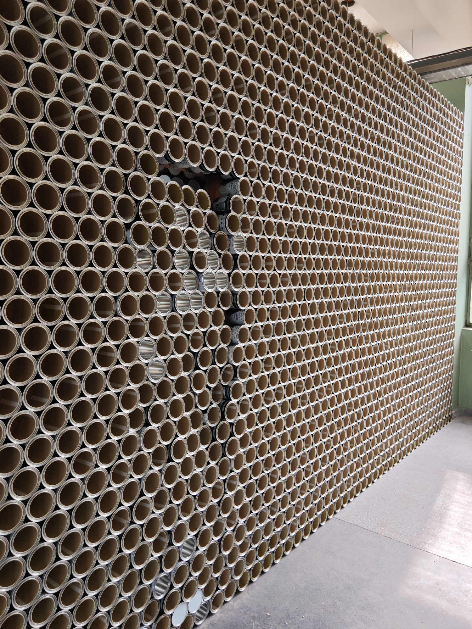This image captures a large, vertically stacked wall of cylindrical containers, occupying the majority of the frame, in what is identified as a facility in Gabrovo, Bulgaria. The containers, which appear to be metal paint cans, are arranged in horizontal rows, with their circular open ends facing the viewer, creating a highly textured, repeating pattern. Each can exhibits a light brown or cardboard-like exterior ring around a metallic, reflective interior.
A prominent feature is a recessed, cross-shaped void or indentation within the stacked wall, located roughly in the center-left. Within this recess, some containers are positioned at a different depth or orientation, revealing more of their silver-colored metallic sides and occasional labels, such as "BLACK & WHITE" and "PAINT" on the left side, and "COAT" on the right.
The floor in the foreground is a light grey concrete or industrial surface. To the right of the stacked containers, a light green wall and a rectangular window or opening are visible in the background. The ceiling, appearing white or light-colored, is partially visible at the top of the frame. The overall environment suggests an industrial storage facility, warehouse, or a large retail setting for paint or similar products. No individuals are present in the scene.