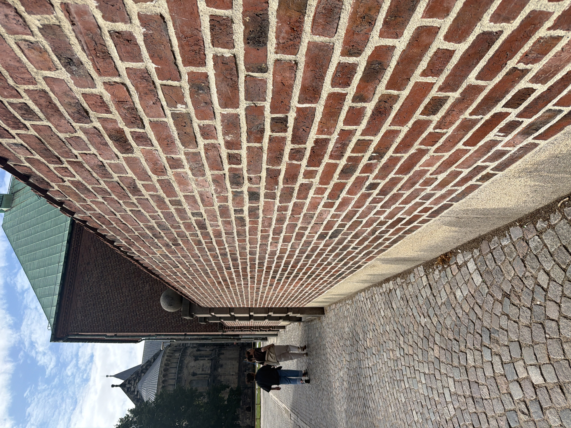 This is a photograph taken at Lund Cathedral in Lund, Sweden. The image shows a cobblestone pathway leading away from the viewer, bordered on the right by a tall, red brick wall. The wall is detailed with visible mortar lines between the bricks. In the distance, on the left side of the pathway, the distinctive architectural features of Lund Cathedral are visible, including its spires and stone facade. Two individuals, appearing to be young women, are walking away from the camera on the cobblestone path, their backs to the viewer. One is wearing a dark top and blue jeans, and the other is wearing a light-colored top and pants. The sky above is blue with scattered clouds. The scene is captured in daylight, suggesting an outdoor setting.