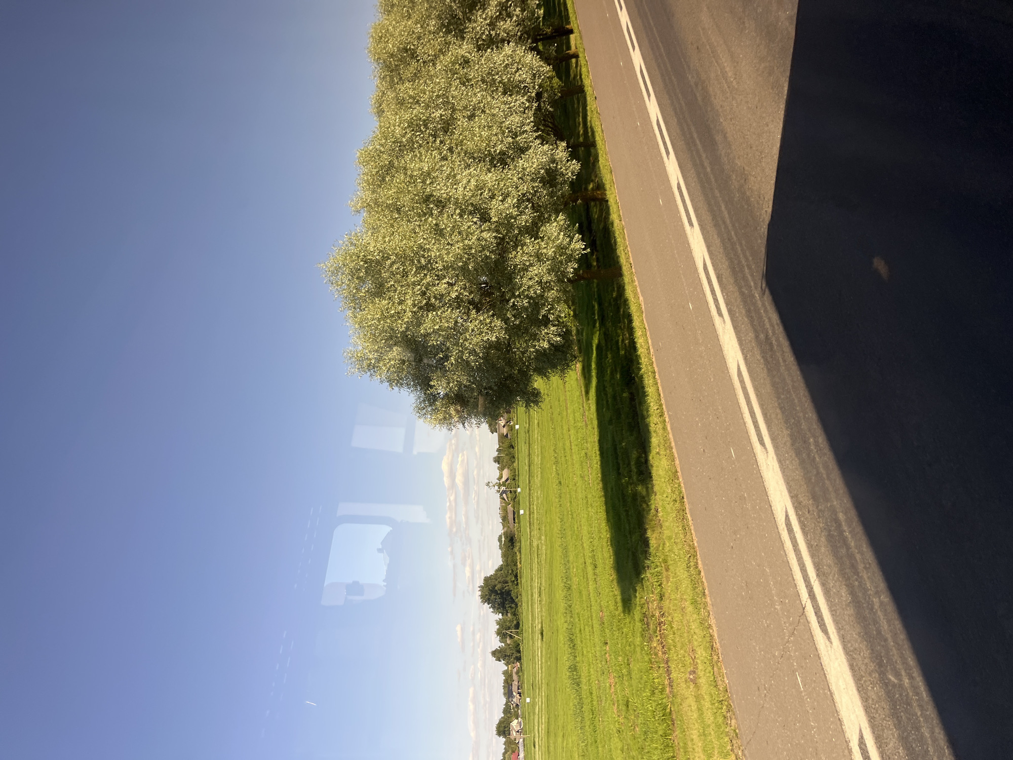 A bright, sunny day is depicted as viewed from a vehicle through a window. The foreground shows a dark asphalt road with white lane markings and a distinct shadow cast by the vehicle. To the left of the road is a vibrant green, mowed field. Beyond the field, a small village or settlement with houses is visible, nestled amongst trees. In the background, the sky is a clear, brilliant blue. The scene appears to be a rural landscape, likely in or near Myadzyel, Belarus, given the characteristic countryside. No people or animals are present.