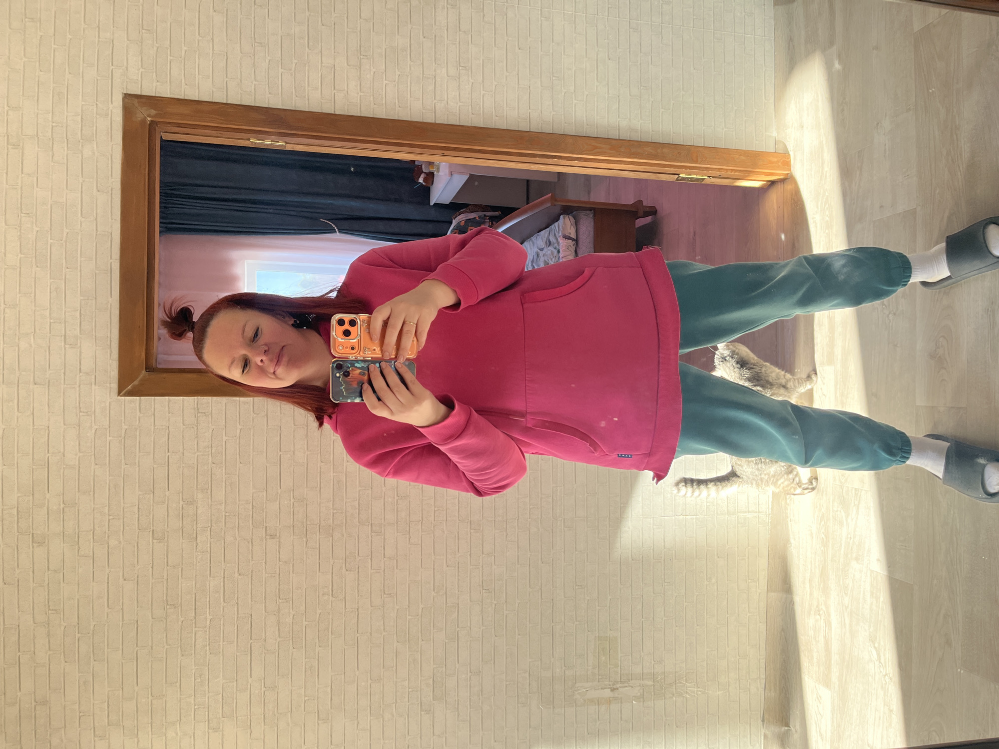 A woman is taking a selfie in a mirror indoors. She is wearing a pink hoodie and teal sweatpants, with white socks and gray slides. She has reddish-brown hair pulled into a small bun. She is holding two phones in her hands, one with a peach-colored case and the other with a dark blue case. The mirror is framed by a wooden doorframe. Behind her in the mirror, a bedroom is visible with a bed and other furniture. A cat is lying on the floor to her right. The background appears to be a wall with a brick pattern. The setting is a room in Chaikovsky, Russia.