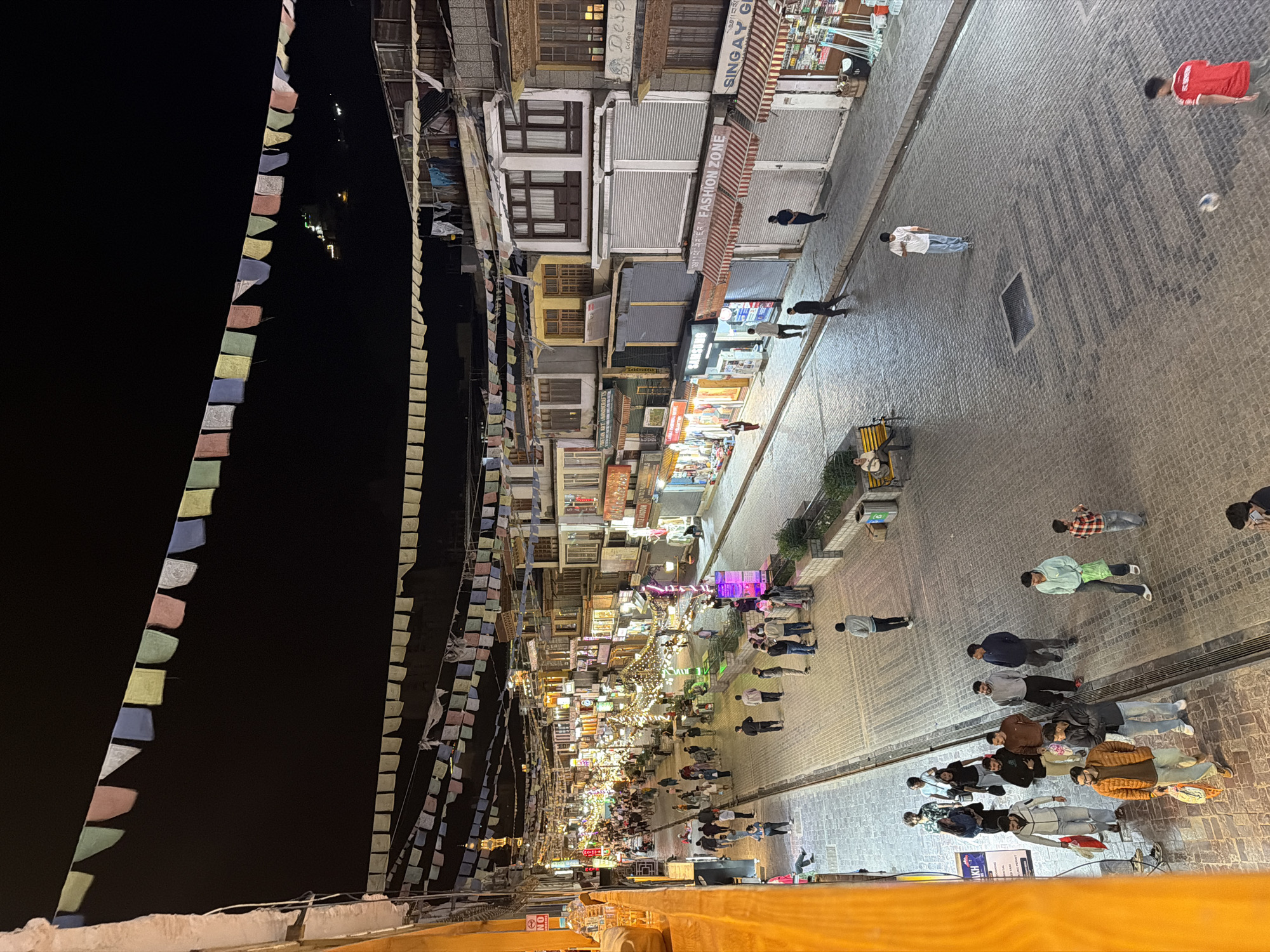 This is a nighttime aerial view of a street in Leh, India, decorated with strings of prayer flags and string lights. The street is paved with cobblestones and lined with multi-story buildings housing shops and businesses. Many shops are illuminated, displaying signs in English and other languages. A crowd of people are walking along the street, some browsing in shops, others conversing. Several individuals are visible in the foreground and middle ground, walking in various directions, some carrying bags. A few people are seated on benches. In the distance, a hill with illuminated structures is visible. The overall scene suggests a lively commercial area during the evening hours.