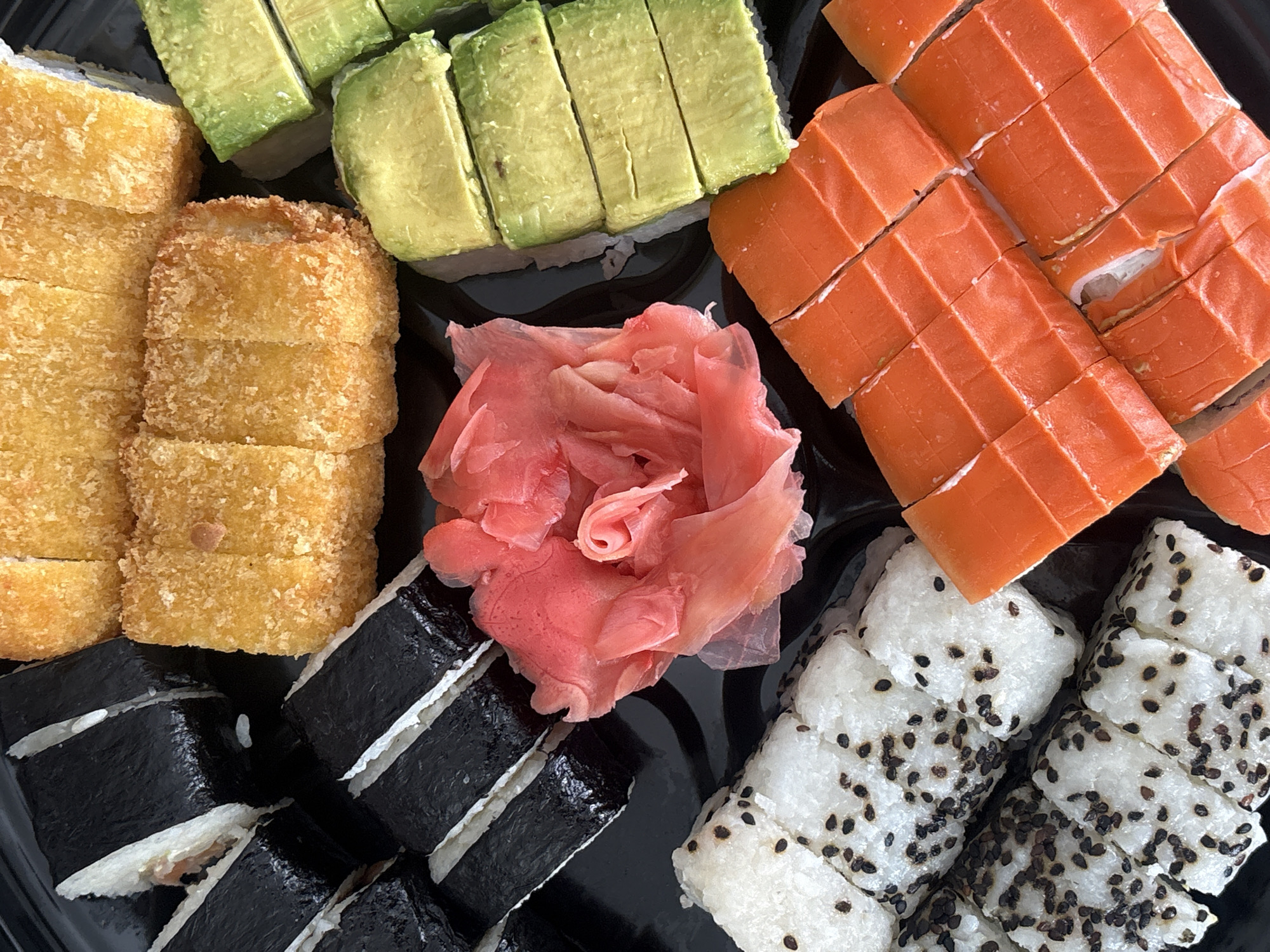 This close-up, overhead shot displays a vibrant assortment of sushi rolls arranged on a black serving tray. The platter features several distinct types of sushi, meticulously cut into individual pieces.
In the upper-left quadrant, there are rolls topped with evenly sliced, bright green avocado. Adjacent to these, on the left side, are golden-brown rolls with a textured, breaded or fried exterior, suggesting a tempura-style preparation. Centrally located, a generous heap of delicate, light pink pickled ginger (gari) provides a pop of contrasting color.
To the right, two rows of sushi rolls are topped with thinly sliced, vibrant orange salmon, cut into rectangular pieces. Below the ginger and extending to the bottom-left, are traditional maki rolls wrapped in dark green nori (seaweed), with glimpses of white rice and filling visible. Finally, in the bottom-right, are rolls with white rice on the outside, generously sprinkled with black sesame seeds, creating a speckled pattern.
The arrangement suggests the sushi is freshly prepared and presented for consumption. The lighting is bright and even, highlighting the textures and colors of the ingredients. There are no people, visible text, or specific location cues within the image itself.