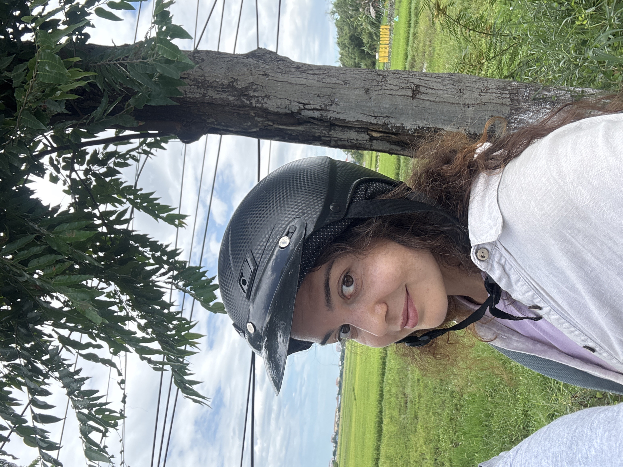 A woman wearing a black helmet and a light-colored open button-up shirt over a purple shirt poses for a selfie. She has brown curly hair and is smiling. She is positioned in front of a tree and a lush green rice field. In the background, there are buildings and a cloudy sky with some overhead power lines. A sign that reads "AN NHIEN GARDEN" is visible in the distance to the right. This scene is likely in Hoi An, Vietnam, a region known for its vibrant green rice paddies and rural landscapes.