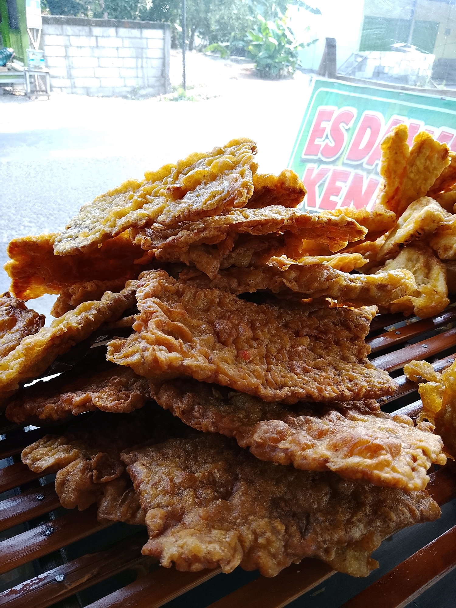 A pile of fried tempeh is displayed on a wooden slatted surface in front of a window. The tempeh pieces are flat and irregular in shape, with a crispy, golden-brown coating. In the background, through the window, a sign with red and white text on a green background is partially visible. The text appears to read "ES DA KENC". Outside the window, a brick wall, some greenery, and a road are visible, suggesting an outdoor setting. The overall scene is likely a food stall or roadside eatery. The location is identified as Banaran, Indonesia. There are no people or animals present in the image.