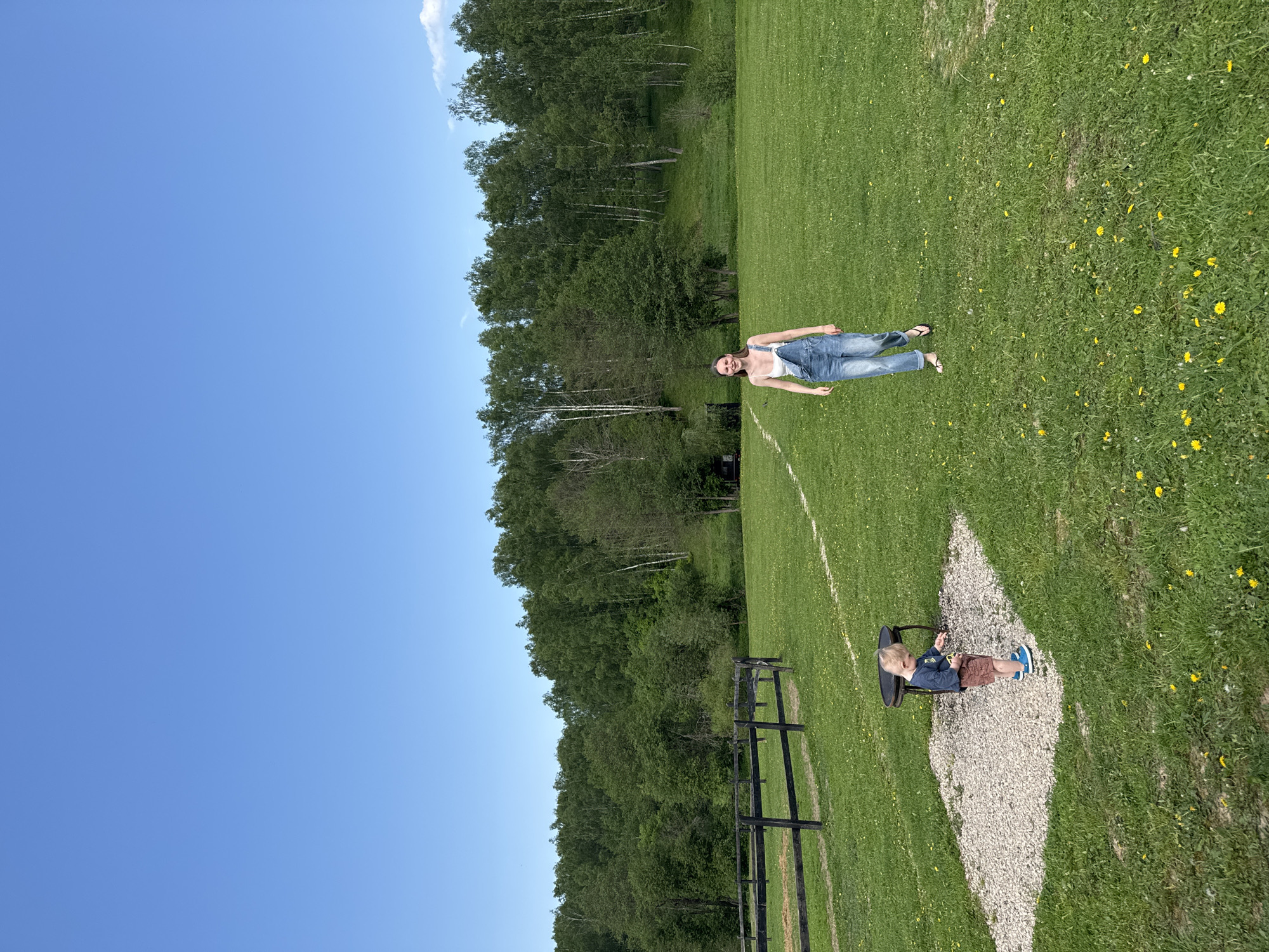 The image shows an outdoor scene in what appears to be a grassy field. In the distance, near a treeline, a woman wearing a white top and jeans overalls is lying on the grass. Closer to the bottom of the frame, a young child is seated in a small chair on a patch of gravel. There is a black wooden fence visible, and the sky is clear and blue. The location is Prudki, Russia.