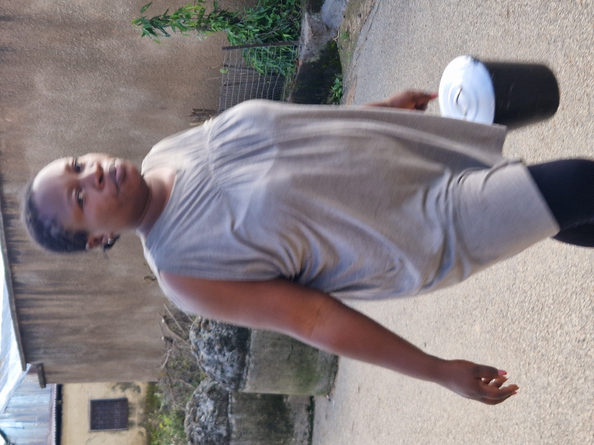 A medium shot captures a Black woman walking outdoors, holding a black bucket with a white lid. She is wearing a sleeveless, grey, gathered-neck dress and dark pants. The woman's face is turned slightly to the left, and her left arm is extended, holding the bucket. In the background, two large, round, grey concrete structures are visible on the left. Behind them is a building with a corrugated metal roof and a tan wall. To the right of the woman, a chain-link fence with some greenery behind it is present. The ground is a paved surface.