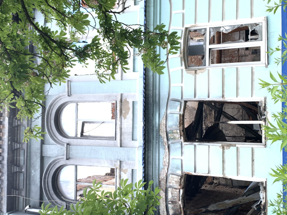 The image shows the facade of a building located at latitude: 49.192, longitude: 37.279 in Izium, Ukraine. The building is light blue with white trim and has five windows. The windows are all broken. The top two windows have their glass panes shattered, while the bottom three windows have been completely blown out. The interior of the building is visible through the broken windows, and there is debris and charred wood inside. The building appears to have been damaged by fire. A tree with green leaves partially obscures the building, but it is growing outside of the structure. The building is likely residential. No people are present.