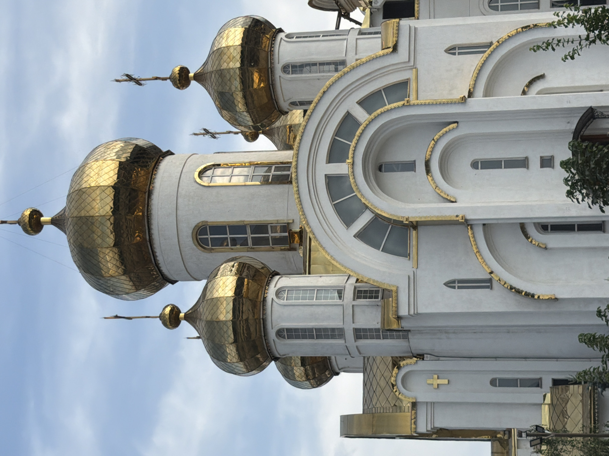 A low-angle, medium shot captures the exterior of a white orthodox church with golden domes under a cloudy sky. The church has a grand, ornate facade with multiple domes of varying sizes, each topped with a cross. The main dome, the largest and highest, is octagonal and covered in gold scales. Smaller domes are visible to its left and right, also adorned with golden scales. The building features numerous arched windows, some with intricate gold detailing around their frames, and a prominent arched entrance with a gold-trimmed awning. A gold cross is affixed to the wall of the church on the left side. Green trees and foliage are visible at the bottom of the frame, partially obscuring the base of the building. The sky is a mix of white clouds and blue.