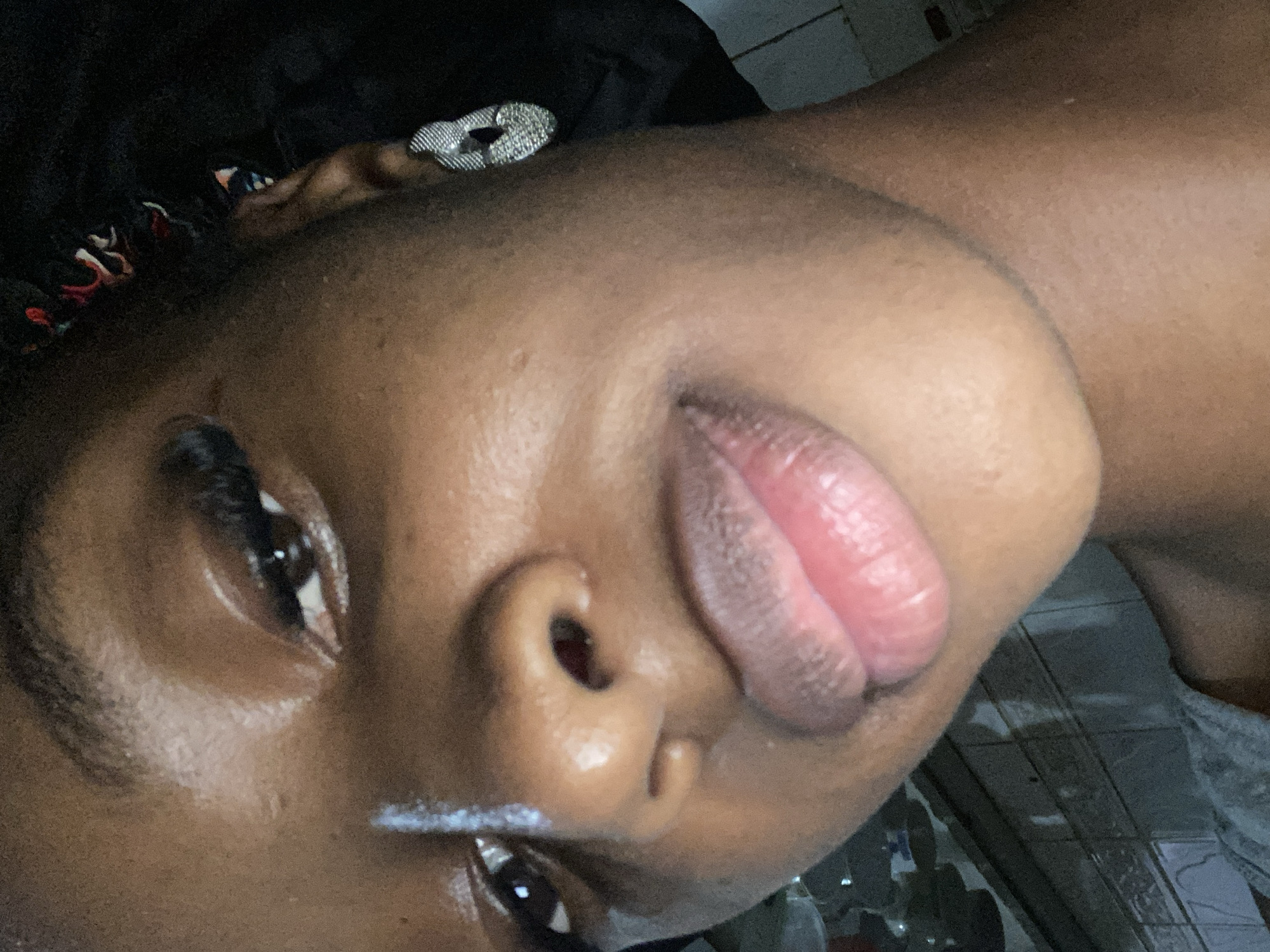 The image is a close-up, low-angle portrait of a young Black woman. She has dark skin, dark eyes, and full lips with a pinkish hue. Her left eye is visible, with long eyelashes. She is wearing a silver, textured hoop earring in her left ear. Her hair is dark and appears to be styled in braids, with colorful fabric woven into it. She is wearing a dark satin bonnet or headwrap. Her skin appears smooth and slightly dewy.
The background is out of focus, but appears to be an indoor setting with tiled walls. The lighting is dim, with some reflections on her skin. There are no discernible landmarks in the image that would definitively place it in Jos, Nigeria, as the background is generic.
