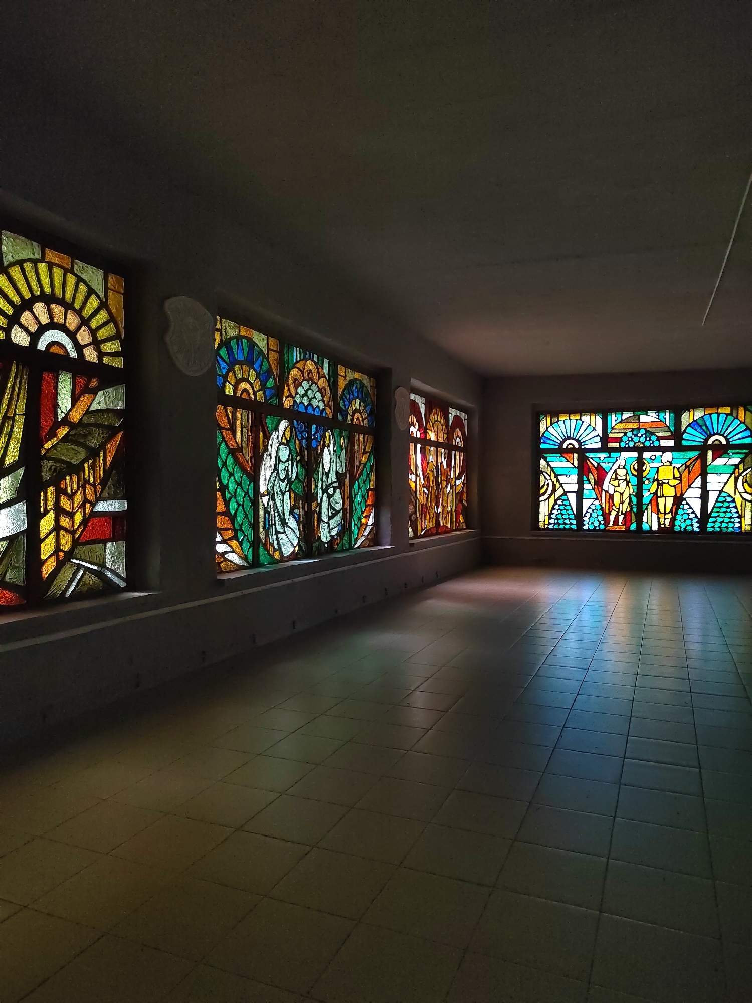 The image shows a long, empty room with tiled flooring. Sunlight streams through large stained-glass windows on the left and right walls, casting colorful patterns on the floor and walls. The windows are adorned with intricate geometric and figurative designs in vibrant hues of yellow, orange, green, blue, and red.
On the left wall, there are three stained-glass panels. The first panel features abstract geometric shapes, predominantly in yellow and orange. The second panel depicts two figures with what appear to be wings, rendered in green and white. The third panel on the same wall also shows figurative elements, possibly people or mythical beings, in warm tones of yellow and orange.
The right wall has a larger stained-glass window with a central design featuring two standing figures in yellow and red, surrounded by fan-like patterns and circular motifs in blue and green. The overall impression is one of solemnity and artistic beauty, likely within a public building or a place of worship, given the nature of the stained glass. The light suggests it is daytime, possibly late afternoon given the long shadows. The location context provided is Свірскі сельскі Савет, Belarus, hinting at a rural or small-town setting within Belarus. There is no visible text in the image.