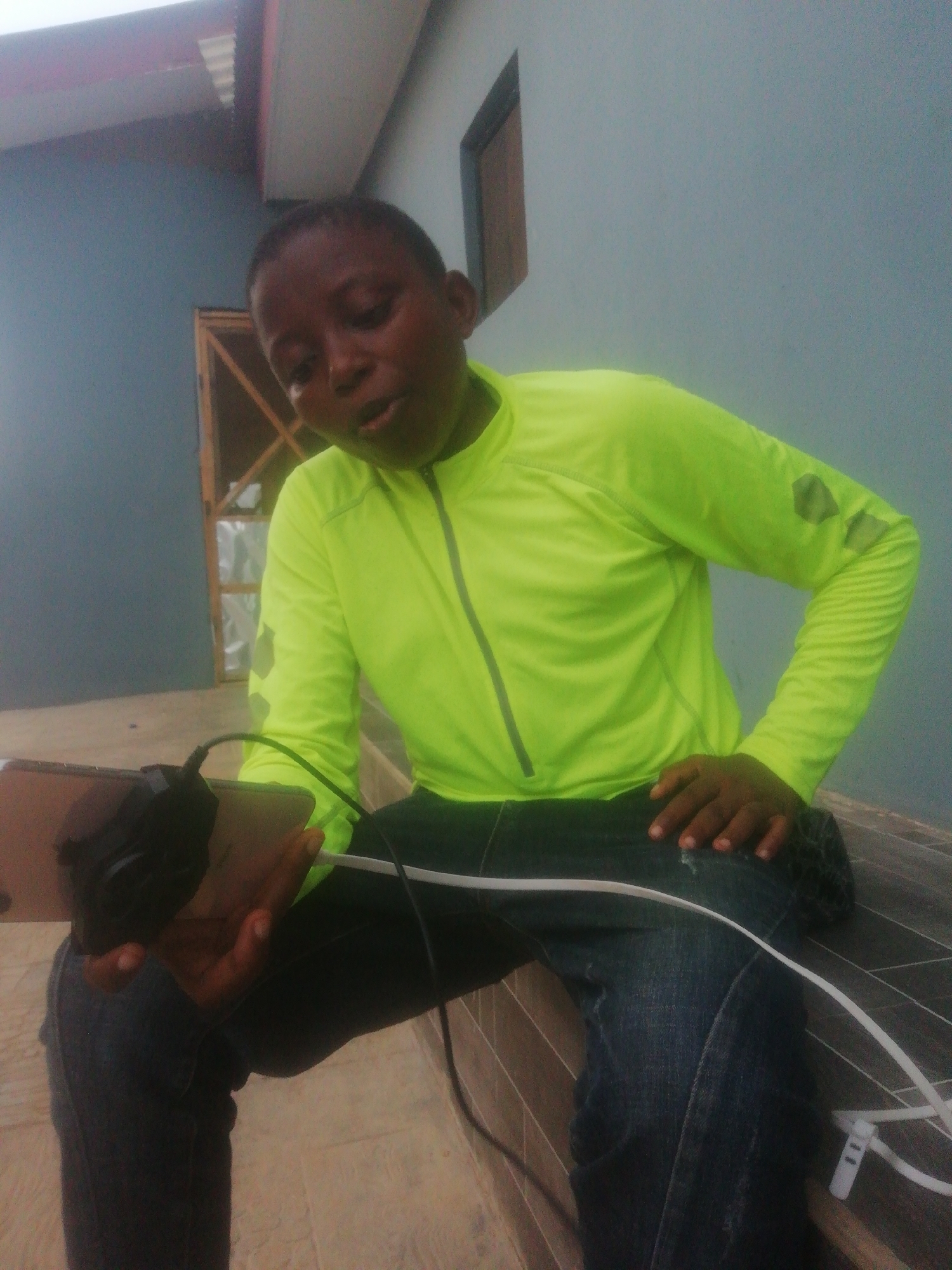 A person is sitting outside on a bench in Ikorodu, Nigeria. The person is wearing a bright green long-sleeved shirt and blue jeans. The person is holding a tablet with headphones attached to it. There are white cables connected to the tablet, which are lying on the bench. Behind the person is a wall and a window.