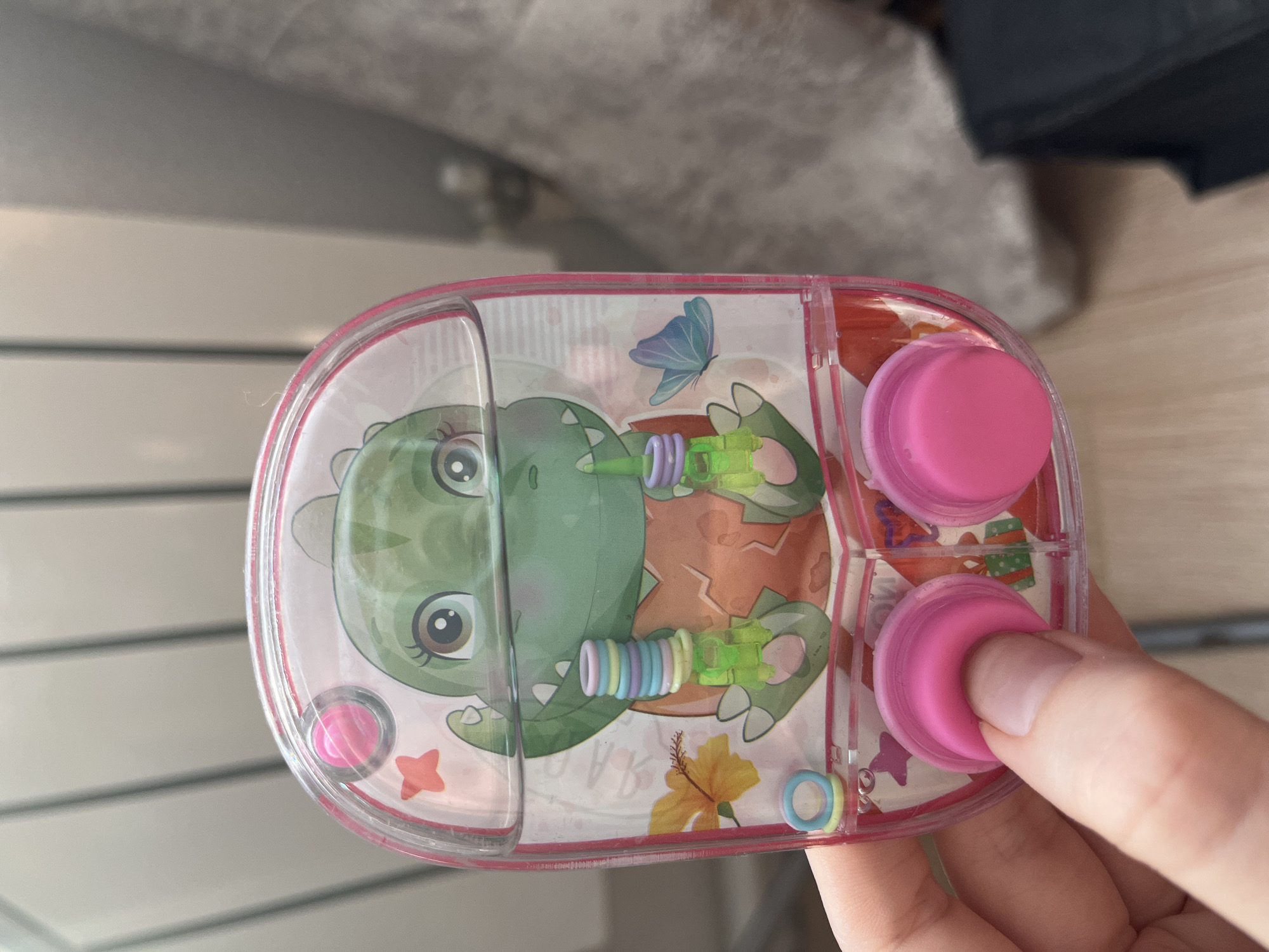 A close-up shot depicts a clear, pink-rimmed water game in the palm of a hand. Inside the game, a cartoon green dinosaur design is visible, along with pink buttons and pastel-colored rings. The background shows a blurred environment. The city of Сосново, Russia is not discernable in this image.