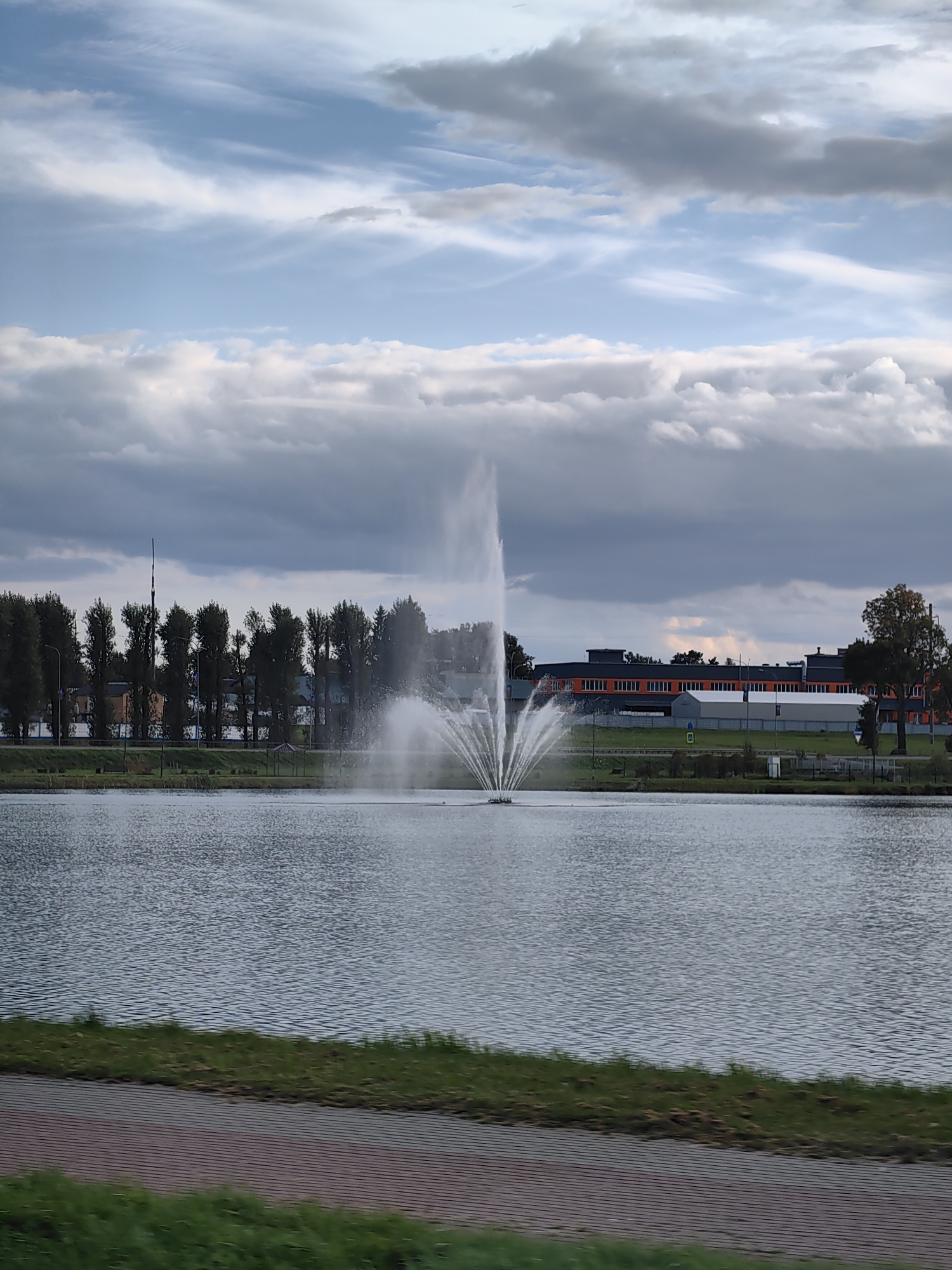 The image depicts a serene lake with a prominent fountain spraying water in the center. The background features a row of trees lining the shore, beyond which are some buildings, including a long structure with orange-trimmed windows. The sky is filled with a mix of light and dark clouds. The foreground shows a pathway or embankment with grass and a patterned surface. The scene appears to be in an urban or suburban setting, likely in the city of Dzyarzhynsk, Belarus, given the context provided. There are no visible people or animals in the image. The overall impression is one of a calm, outdoor environment with a touch of man-made landscaping.