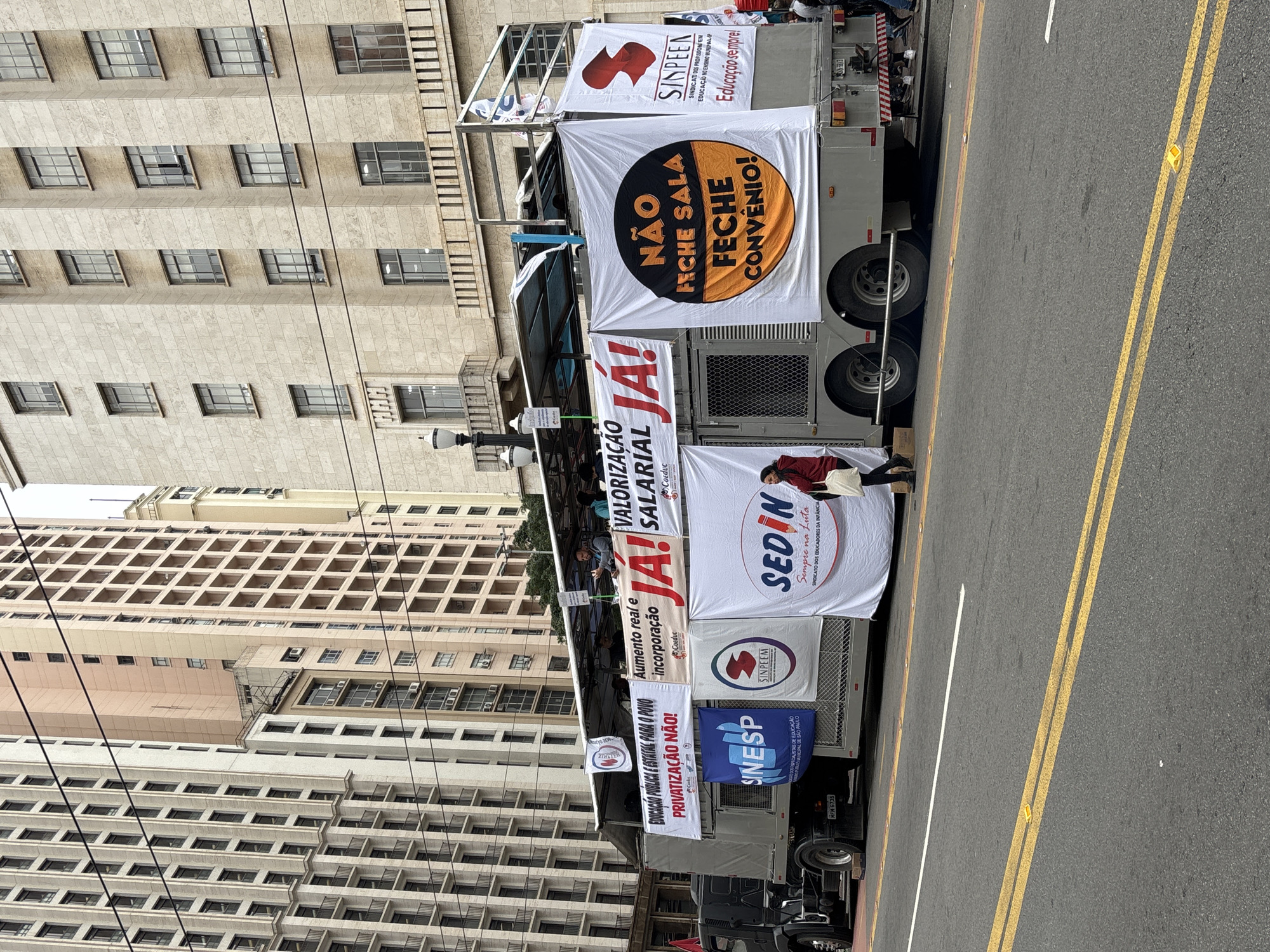 Here is a description of the image:
The image shows a political demonstration or protest in the city of São Paulo, Brazil. A truck is parked on a street with yellow lane markings, and is covered in banners and signs with Portuguese slogans and logos of unions or organizations. Some signs reference "PRIVATIZAÇÃO NÃO" ("No Privatization"), "VALORIZAÇÃO SALARIAL" ("Salary Appreciation"), and "NÃO FECHE SALA, FECHE CONVÊNIO!" ("Don't Close Classrooms, Close Agreements"). The buildings in the background are tall and appear to be made of light-colored stone or concrete. A person in a red jacket is standing next to the truck.