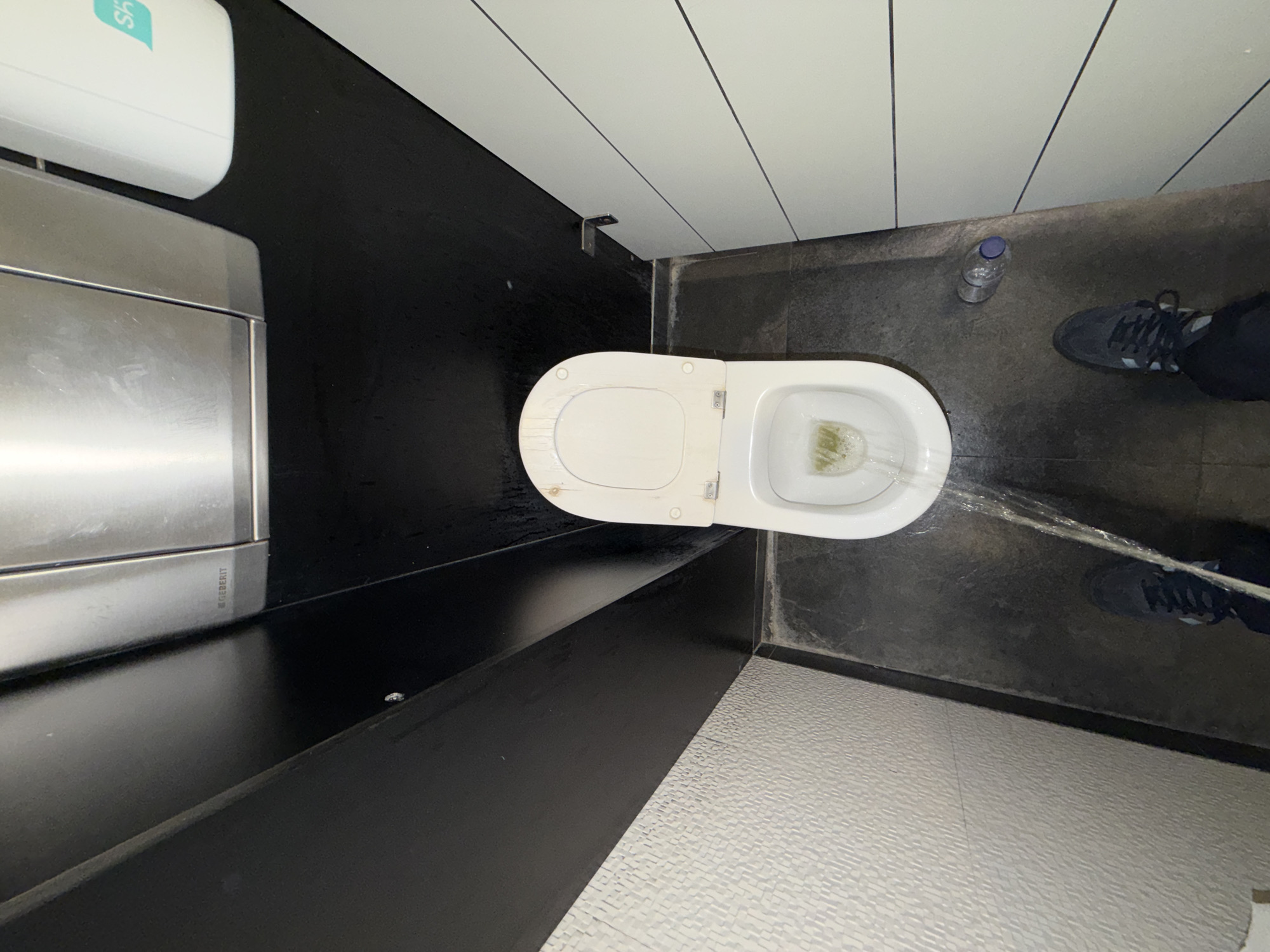 The scene is a top-down view of a restroom. A white toilet is positioned on the right side, lid open. A bottle of water is on the floor. A person is visible by the feet in black trousers and grey shoes. Water is flowing out of the toilet, towards the person's feet. On the left, there is a grey metal sink with a white soap dispenser above. The surrounding surfaces are a mix of black and white materials. The city and country are given as Porto, Portugal.