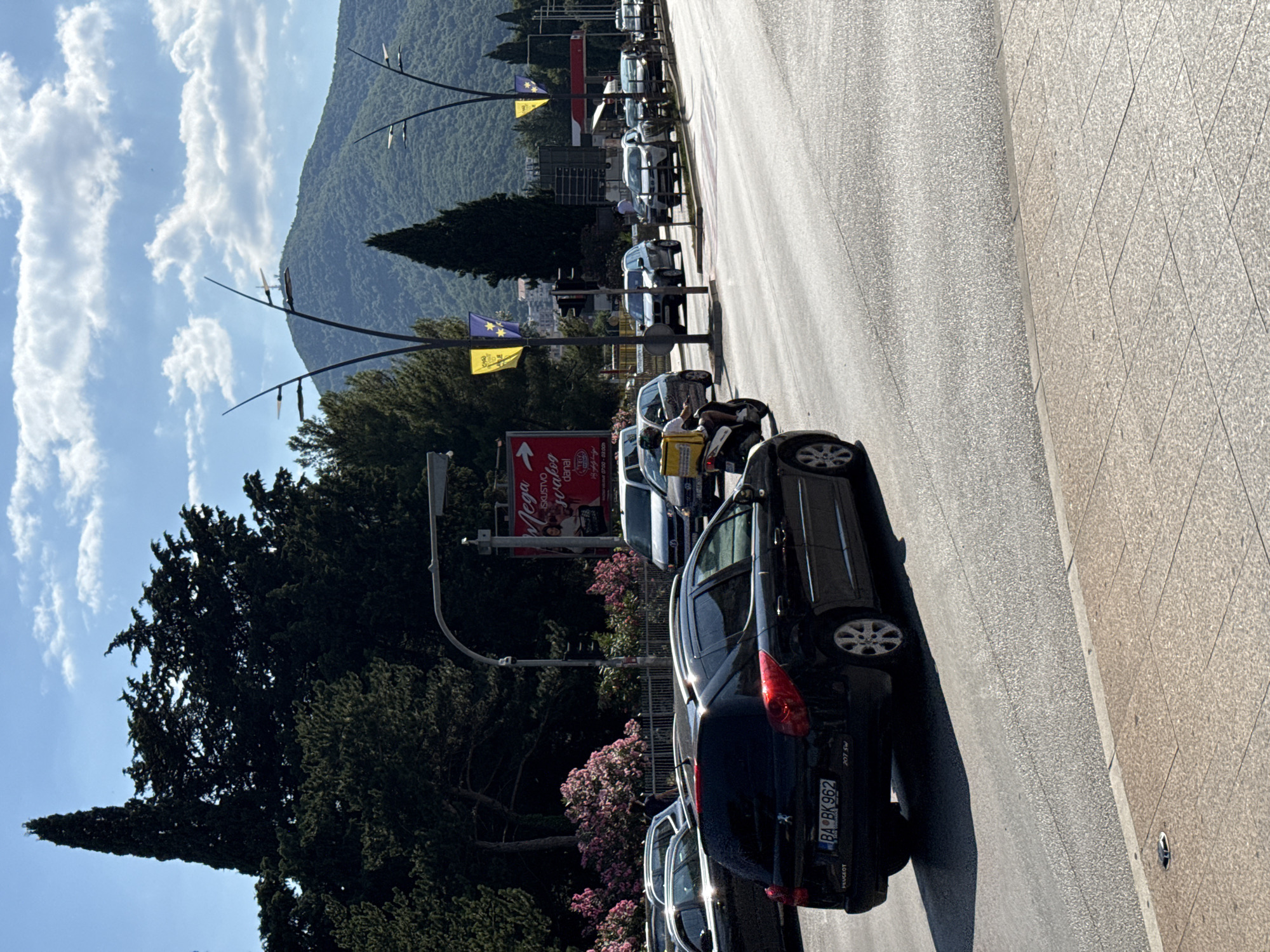 A black Peugeot 207 SW with license plate BA BK 962 is driving on a paved road in Budva, Montenegro. The car is in the foreground, angled slightly towards the right. Behind the car, there is traffic congestion with several other vehicles visible. A person on a scooter with a yellow delivery box on the back is also present. In the background, there are trees, a mountain, and a blue sky with white clouds. A large sign with red and white lettering is visible on the left side of the road. The scene appears to be a street in a city, possibly near a commercial area.