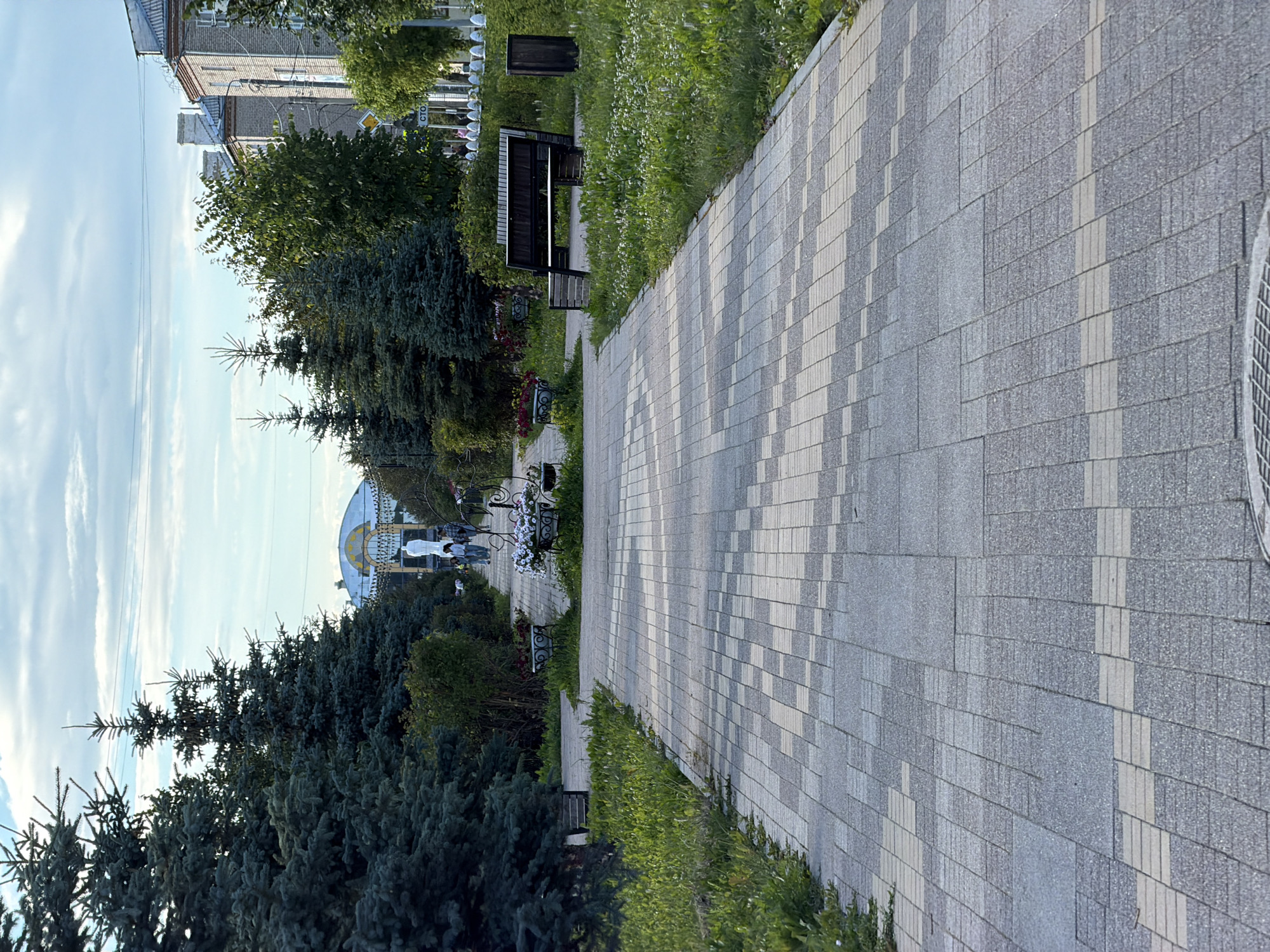 The image depicts a paved walkway in a park-like setting with lush green trees and manicured bushes lining the path. In the distance, a few individuals are walking towards a large, ornate structure with a domed roof. The sky above is partly cloudy. To the right of the walkway, there is a wooden bench and a trash receptacle, surrounded by green grass with small white flowers. The walkway itself is paved with a pattern of light and dark grey rectangular tiles. The overall scene suggests a peaceful outdoor public space.