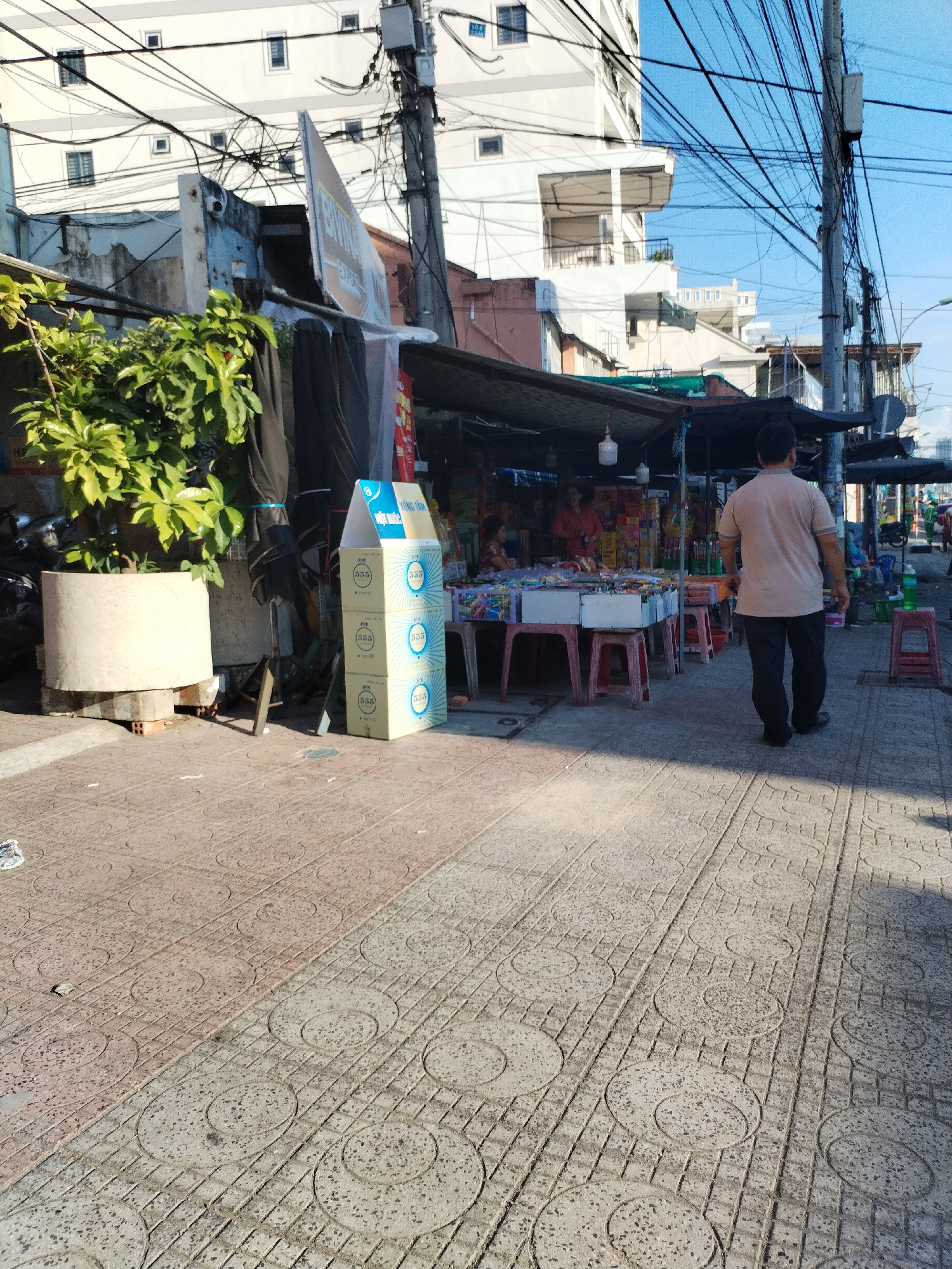 A man walks on a patterned, paved sidewalk in Bac Nha Trang Ward, Vietnam, past a small roadside shop under a makeshift awning. The shop displays an assortment of goods on tables, with two people standing behind the tables. Next to the sidewalk, there is a potted plant, three stacked boxes, and electrical wires overhead. Tall buildings stand in the background.