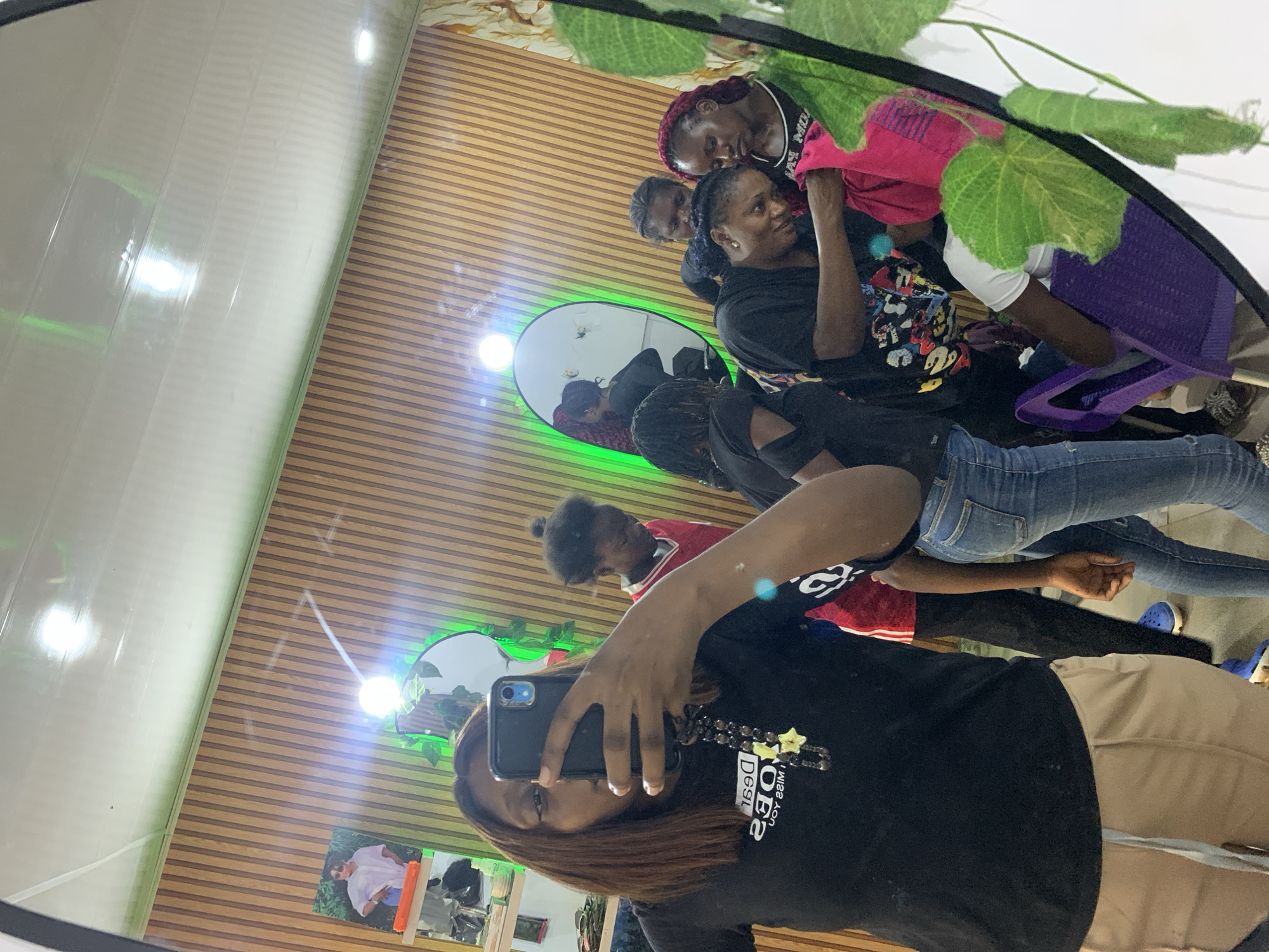 A mirror selfie in a room with a horizontally-paneled wall. A woman holds a phone, facing the mirror, wearing a black t-shirt and khaki pants. Reflected in the mirror are other people. The mirror and the wall are decorated with artificial plants, and the scene suggests a possible location within a city of Jos, Nigeria.