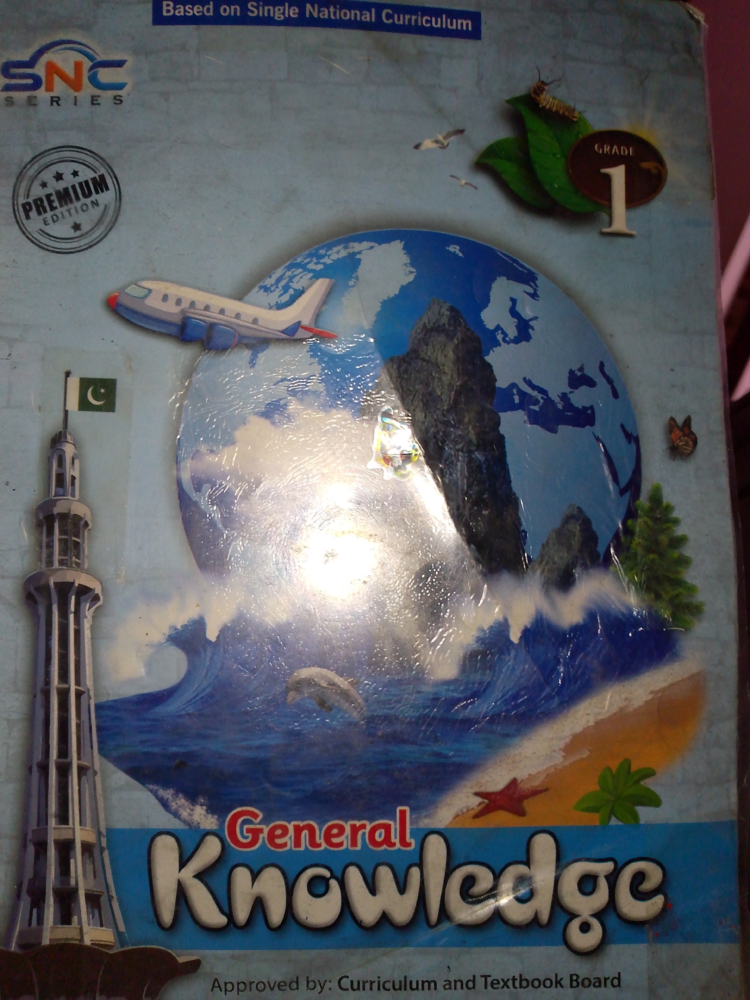 The image is a close-up of a book cover titled "General Knowledge" for Grade 1. The cover features a colorful illustration of a globe with various elements such as an airplane, a minaret with a Pakistani flag, a crashing wave, a dolphin, a starfish, a small tree, a caterpillar on a leaf, and a butterfly. The background is a light blue sky with two birds flying. The book also has "SNC SERIES" and "PREMIUM EDITION" printed on it, and the text "Based on Single National Curriculum" at the top. The bottom of the cover shows "Approved by: Curriculum and Textbook Board." There is no information in the image that suggests a specific location within a city or any people.