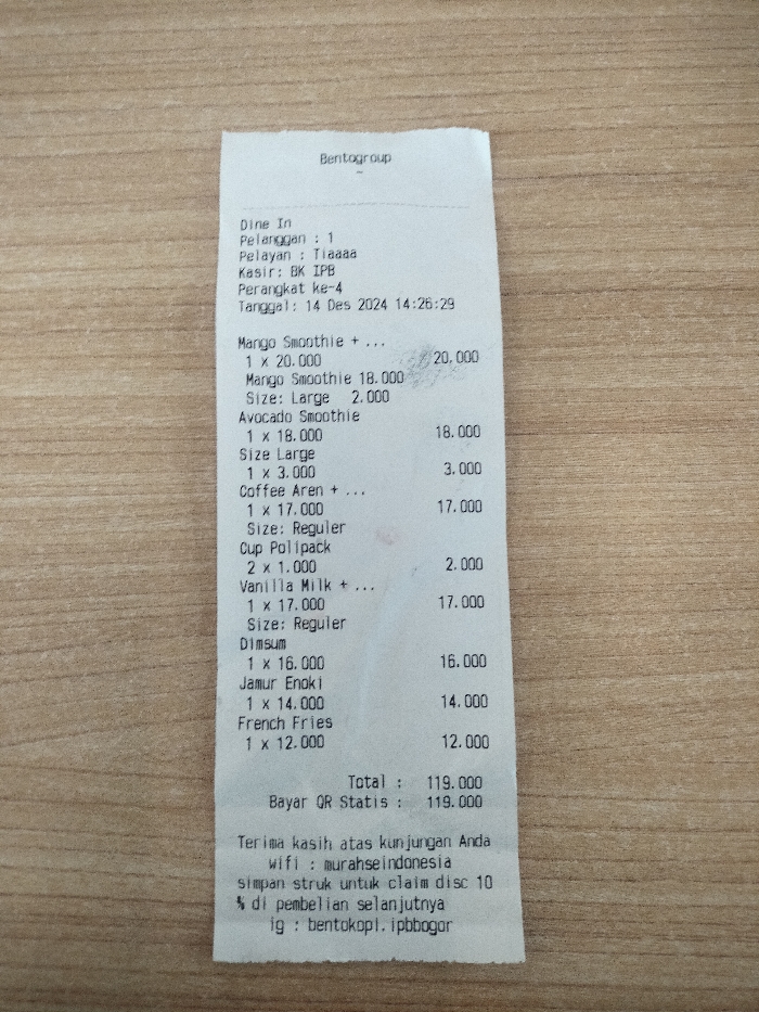 A white paper receipt from "Bentogroup" is centered on a light brown wooden surface, likely a table, in a close-up shot. The receipt, slightly curled at the top, details a "Dine In" transaction for one customer, processed by cashier "BK IPB" on December 14, 2024, at 14:26:29.
The itemized list includes:
- One Mango Smoothie (likely large) for 20,000 (IDR).
- One Avocado Smoothie for 18,000 (IDR), with an additional 3,000 (IDR) for its large size.
- One regular Coffee Aren for 17,000 (IDR).
- Two Cup Polipack for 2,000 (IDR).
- One regular Vanilla Milk for 17,000 (IDR).
- One Dimsum for 16,000 (IDR).
- One Jamur Enoki for 14,000 (IDR).
- One French Fries for 12,000 (IDR).
The total amount is 119,000 (IDR), paid via "QR Statis." The receipt concludes with a thank you message, a Wi-Fi password "murahseindonesia," an offer to claim a 10% discount on the next purchase by saving the receipt, and the Instagram handle "bentokopi.ipbbogor," indicating a location near IPB University in Dramaga, Indonesia.