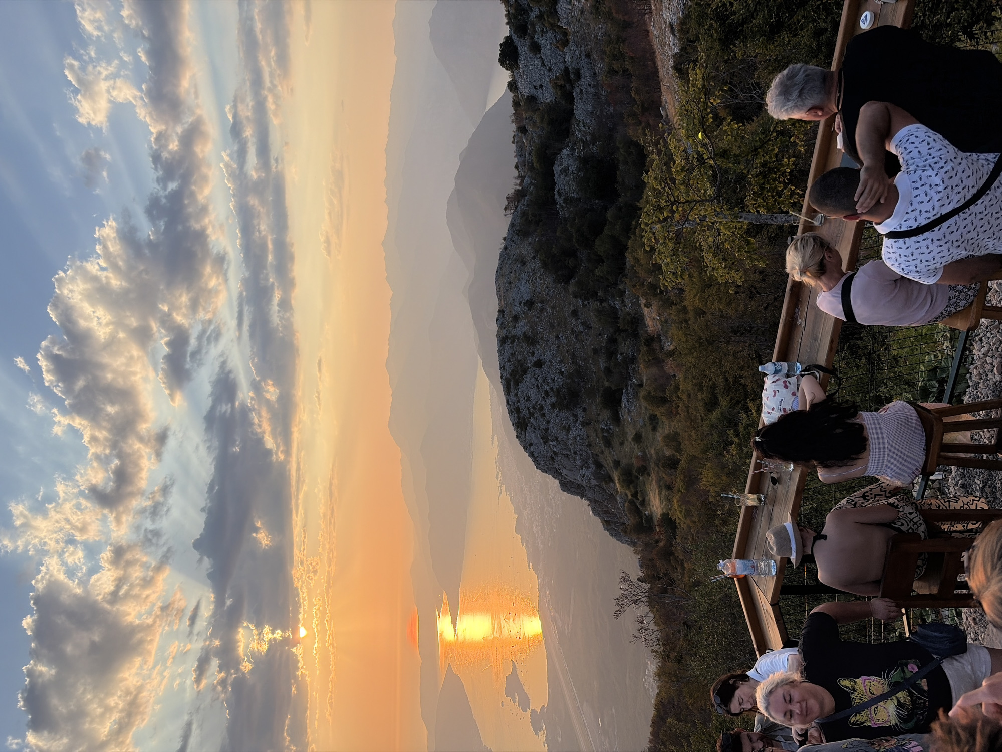 An outdoor bar scene is depicted at sunset, overlooking a body of water and a mountainous landscape. The sun, low on the horizon, casts a golden glow across the sky and reflects on the water. Several people are visible at the bar, some seated and others standing. They appear to be enjoying drinks and the view. The bar itself is made of wood, with stools and drinks on display. The surrounding environment features rocky terrain and scattered trees, indicative of a mountainous or hilly region. The location is identified as a bar in Kuk, Montenegro.