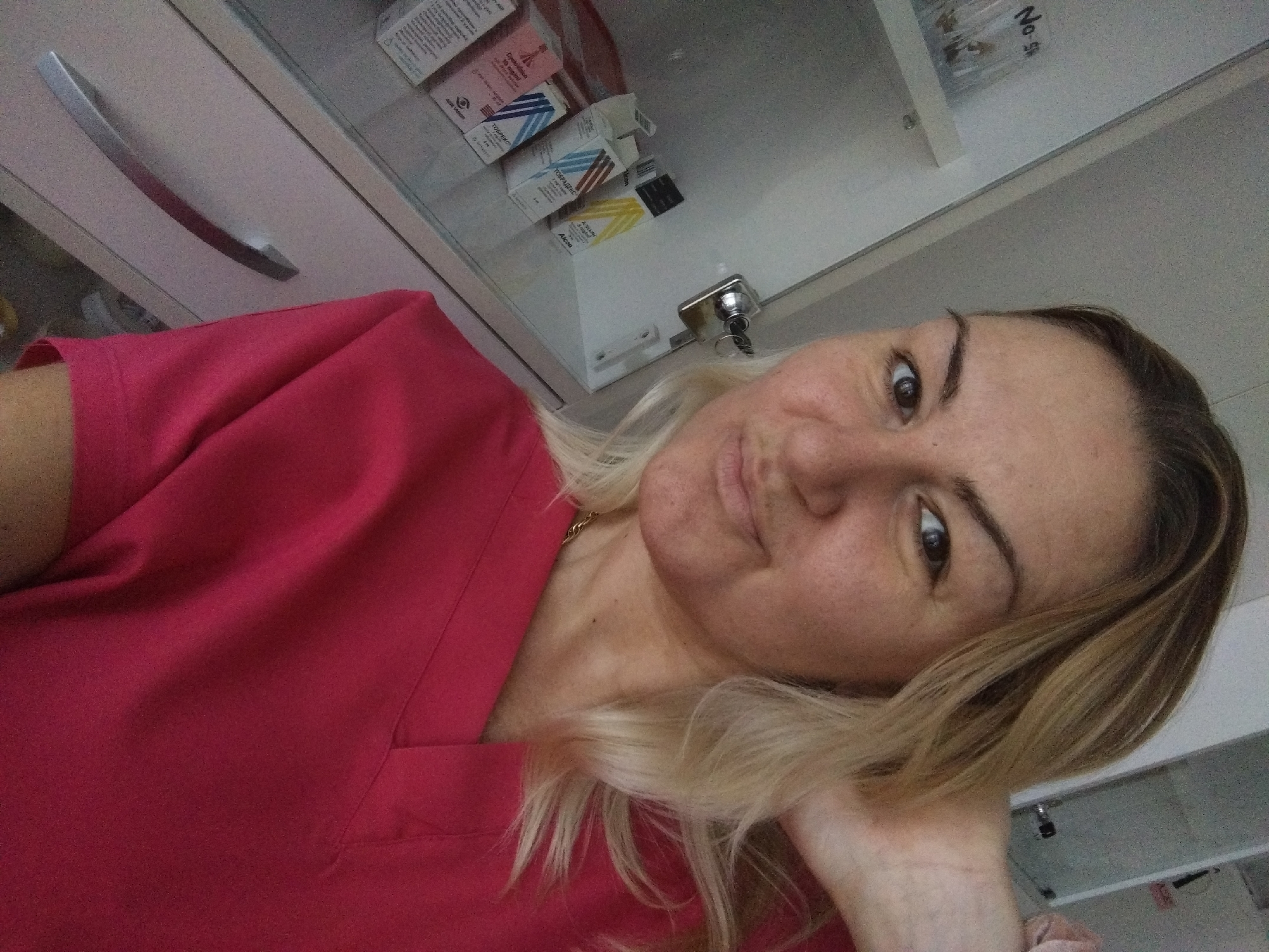 A woman with blonde hair and a pink top is posing for a selfie indoors. She has brown eyes and is resting her head on her hand. Behind her, a cabinet with a silver handle and glass doors contains boxes of medications, labeled with Cyrillic text. A key is in the cabinet lock. A sign saying "No 5" is visible on the upper part of the cabinet. The location appears to be an office or clinic, possibly in Stara Zagora, Bulgaria, given the language on the medication boxes. The focus is on the woman, with the background providing context.