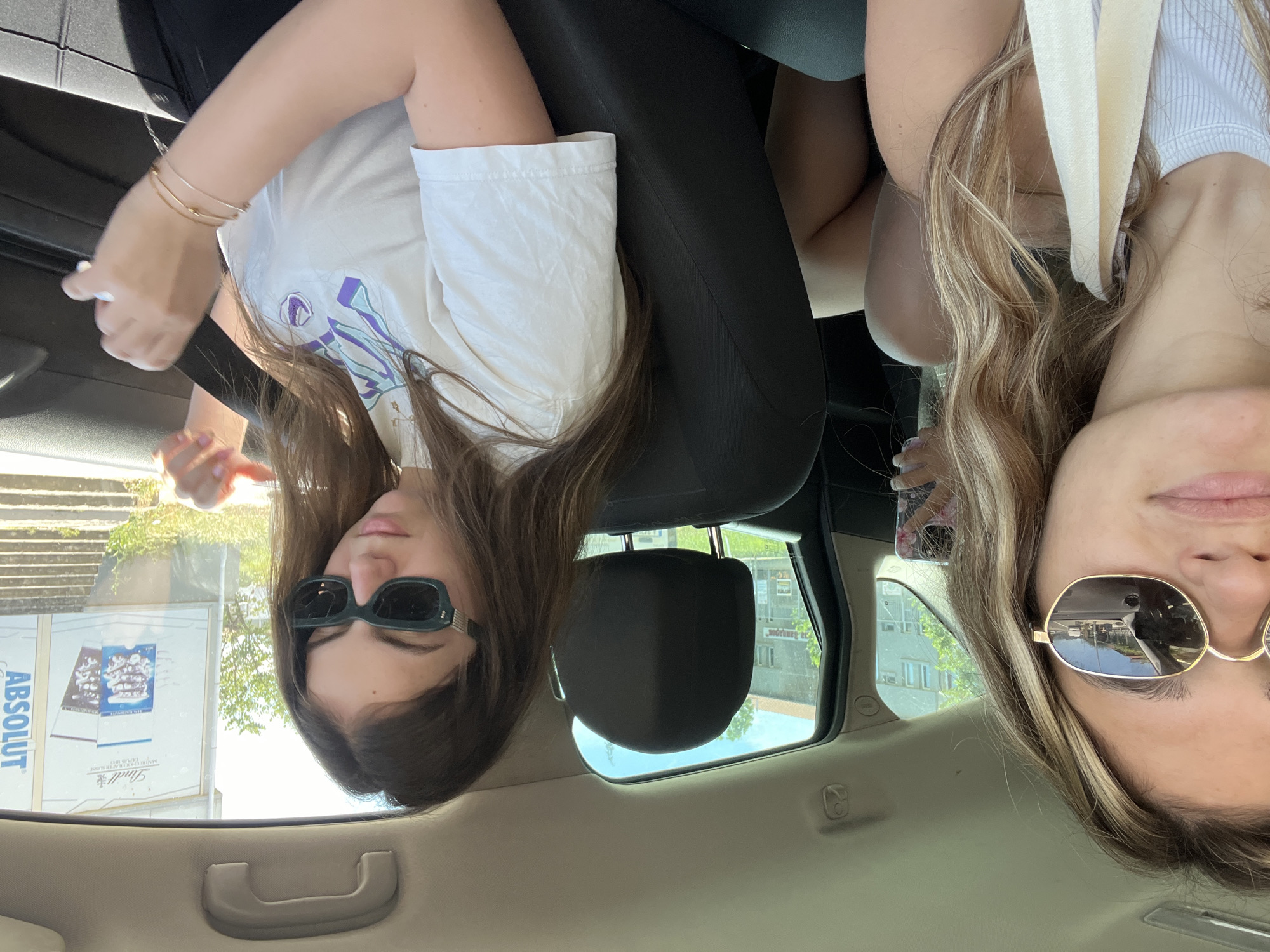 The image shows two young women in a car, with their faces close to the camera. The photo is taken from inside the car, and the perspective is from the backseat. One woman is on the left, wearing a white t-shirt and dark sunglasses. She has long, dark hair and is looking down. On the right is another woman with blonde hair, wearing a white top. She is also looking towards the camera, and her face is partially obscured by a beige strap. The car's interior is visible, including car seats and a handle on the door. Outside the car windows, trees and buildings can be seen, suggesting an urban or suburban environment. There are advertisements on a building in the background, one of which displays the text "ABSOLUT" and "Lindt". The lighting suggests it is daytime, with bright sunlight. The overall tone of the image seems casual and candid.