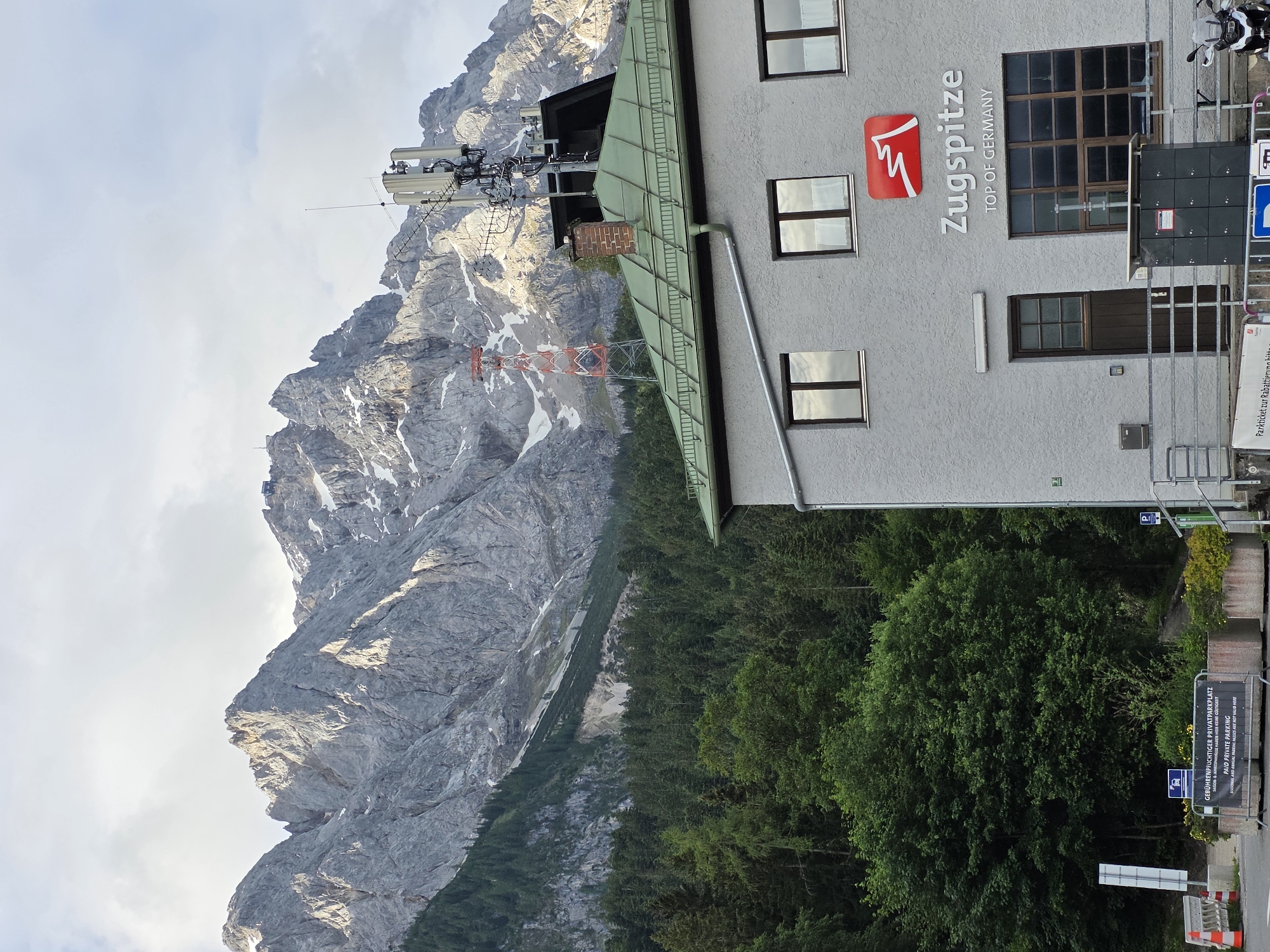 This is a full outdoor, medium shot of the Zugspitze building in Grainau, Germany, with the Zugspitze mountain range in the background. The building is a light gray, two-story structure with a green roof. The Zugspitze logo, a red mountain silhouette with the text "Zugspitze TOP OF GERMANY" below it, is displayed on the building's facade. Several windows are visible, some reflecting the cloudy sky.
A large telecommunications tower with multiple antennas stands on the roof of the building, and a red and white lattice tower is visible on the mountain slope in the distance. The mountain range is rugged and rocky, with patches of snow. A dense forest of dark green trees covers the lower slopes of the mountains.
In the foreground, there is a paved area with parking signs and barriers. A silver motorcycle is parked on the right side of the image. The sky is overcast with scattered clouds. The scene is quiet and appears to be early in the day or late in the afternoon, given the lighting.