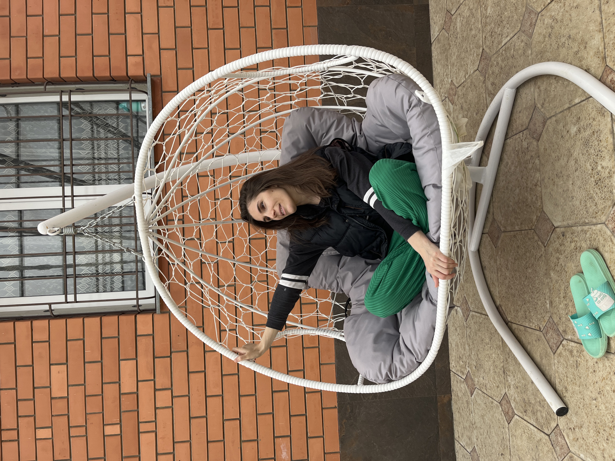 A young woman is sitting in a white hanging egg chair with gray cushions, located outside against a brick wall and stone-tiled ground. She is wearing a black vest, a long-sleeved black shirt with white stripes on the arm, and green pants. Her dark hair falls to her shoulders. Her left hand is touching the side of the chair, and she is smiling. The location is possibly Kaspiysk, Russia. A window with decorative bars is partially visible above the brick wall. A pair of light green sandals is on the ground near the base of the chair.