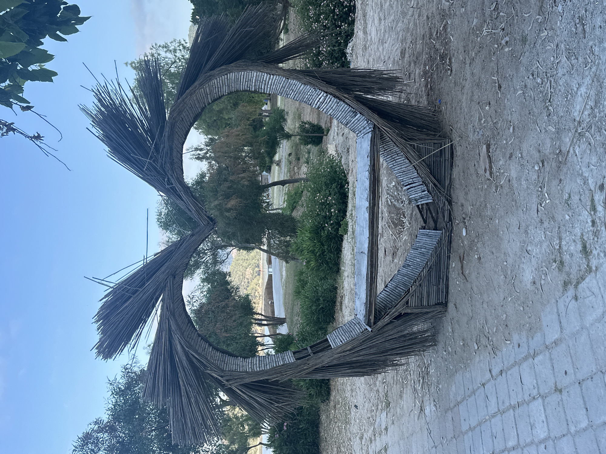 The image displays a large, heart-shaped decorative structure situated on sandy ground in Fethiye, Turkey. The structure is constructed from numerous dark, thin wooden sticks or dried palm fronds, intricately woven around a core frame composed of lighter, horizontally segmented bamboo pieces that define the heart's curvature. Many of the dark sticks protrude outwards, particularly at the top, forming a spiky texture. To the lower left, a paved walkway made of light-colored square blocks abuts the sandy area. The ground around the structure is sandy, showing sparse dry vegetation and scattered organic debris. In the mid-ground, a dense arrangement of green trees of varying sizes extends towards the background. A body of water, likely a bay, with a sandy shoreline is visible between the trees in the far background, with distant hills beneath a clear blue sky. No individuals, animals, or specific actions are depicted in the scene, which is illuminated by natural daylight.