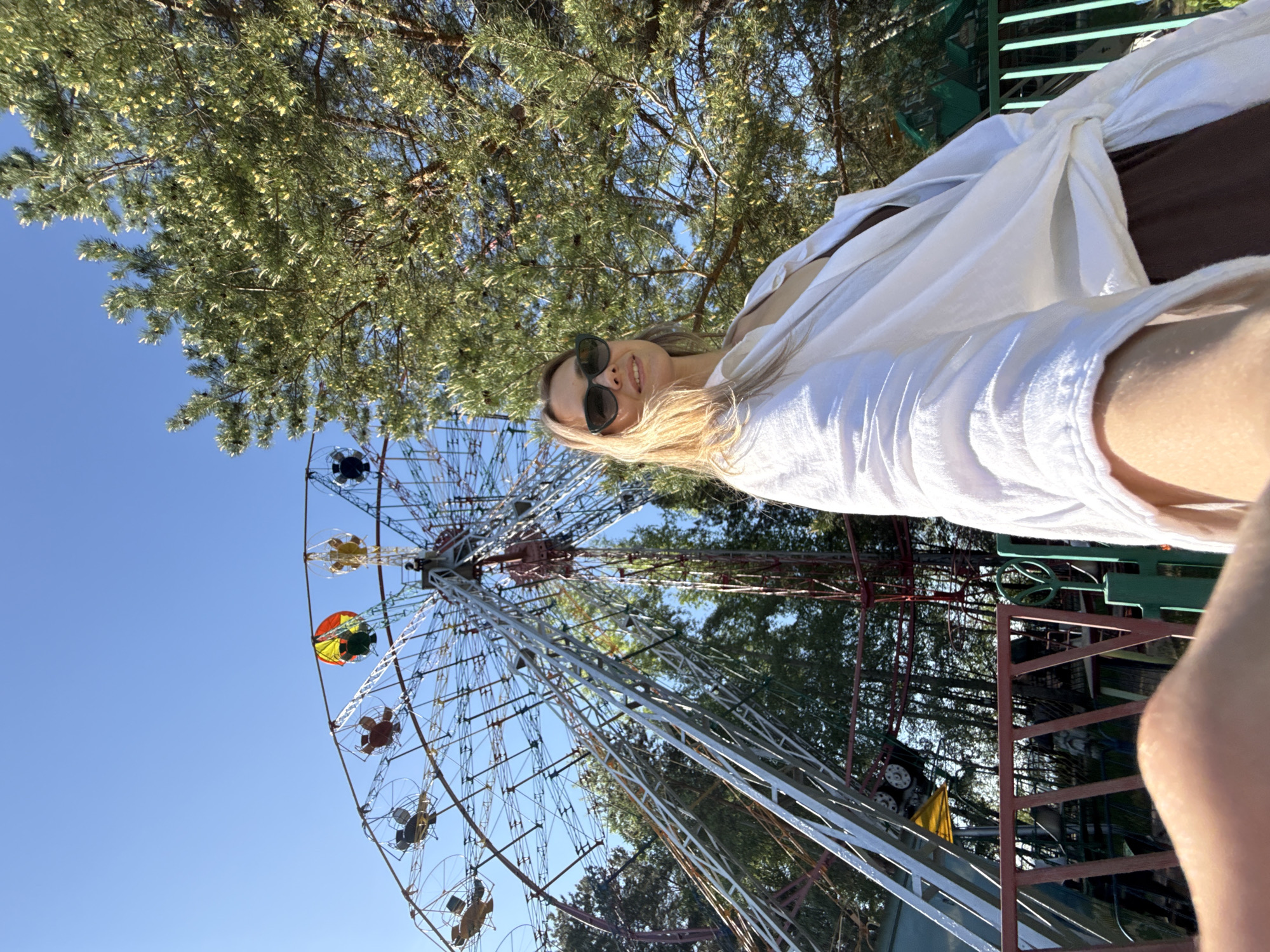 A woman with blonde hair and sunglasses is posing for a selfie in front of a Ferris wheel in Chaikovsky, Russia. She is wearing a white shirt and brown pants. The Ferris wheel is a tall, metal structure with brightly colored seats. Trees surround the Ferris wheel. The sky is blue and sunny.