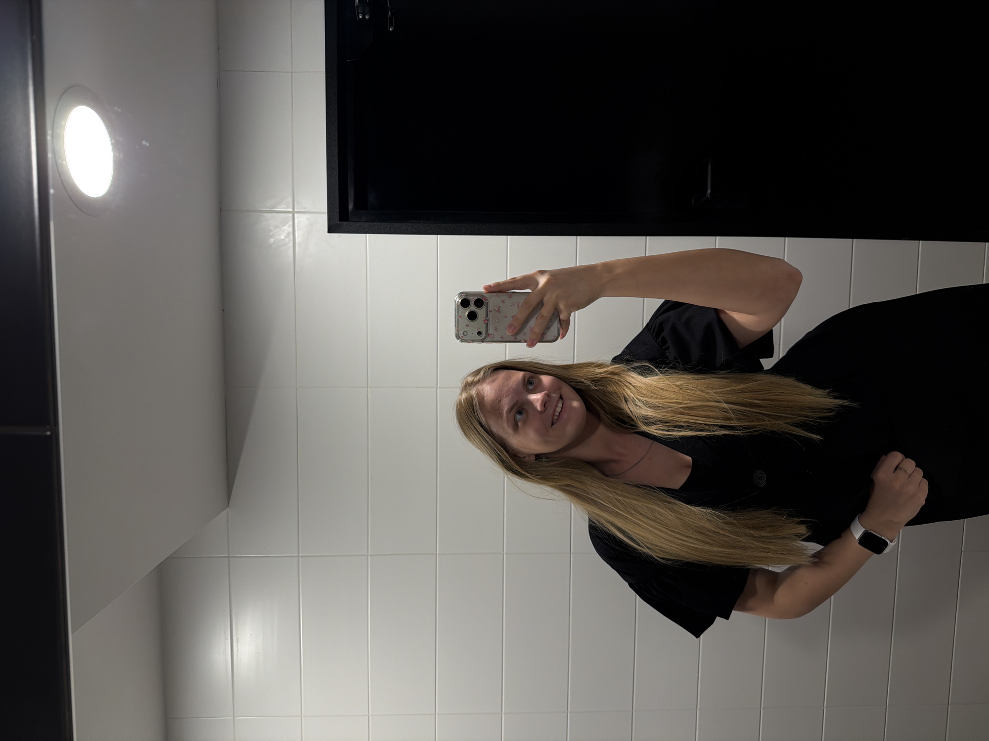 A young woman stands in a bathroom in Izhevsk, Russia, taking a selfie in front of a tiled wall with a black cabinet. She is wearing a black dress and a white smartwatch. Her blonde hair is long and loose. The bathroom has white tiles and a round, recessed light in the ceiling. The black cabinet above her is rectangular.