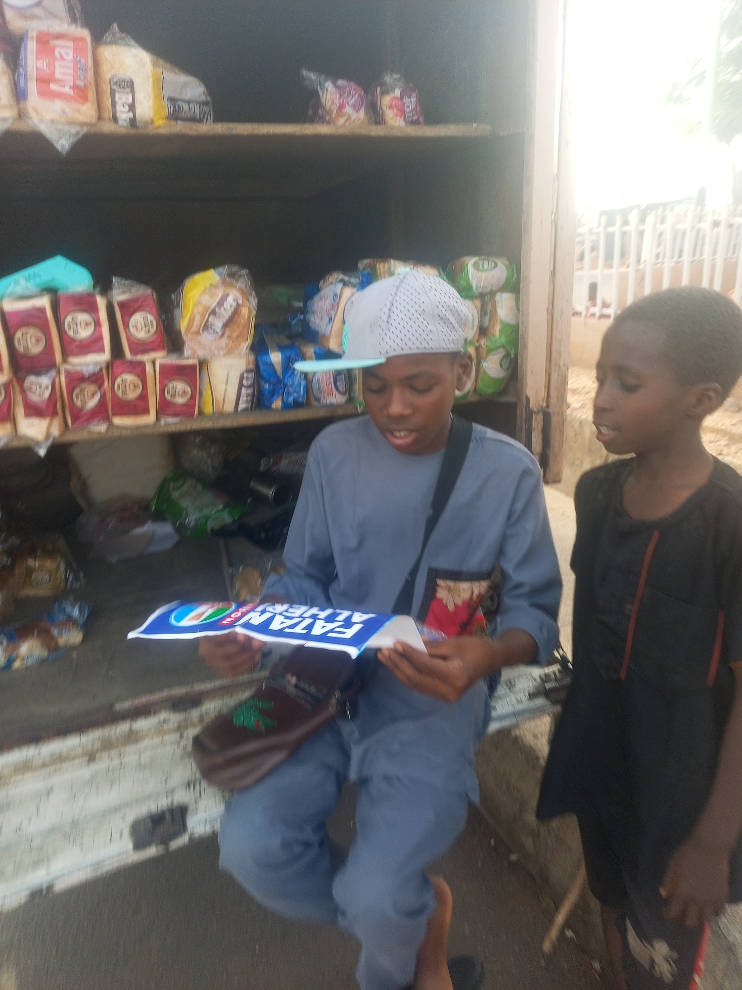 In an outdoor setting, two boys stand beside the back of a truck transformed into a makeshift bread shop. The truck's open rear reveals shelves stocked with various packaged loaves of bread. The boy on the left, wearing a gray cap and matching attire with a satchel, intently examines a blue and white document, possibly promotional material. The boy to his right, dressed in black, looks towards the first boy, seemingly observing. The location appears to be in the city of Unknown, Unknown.