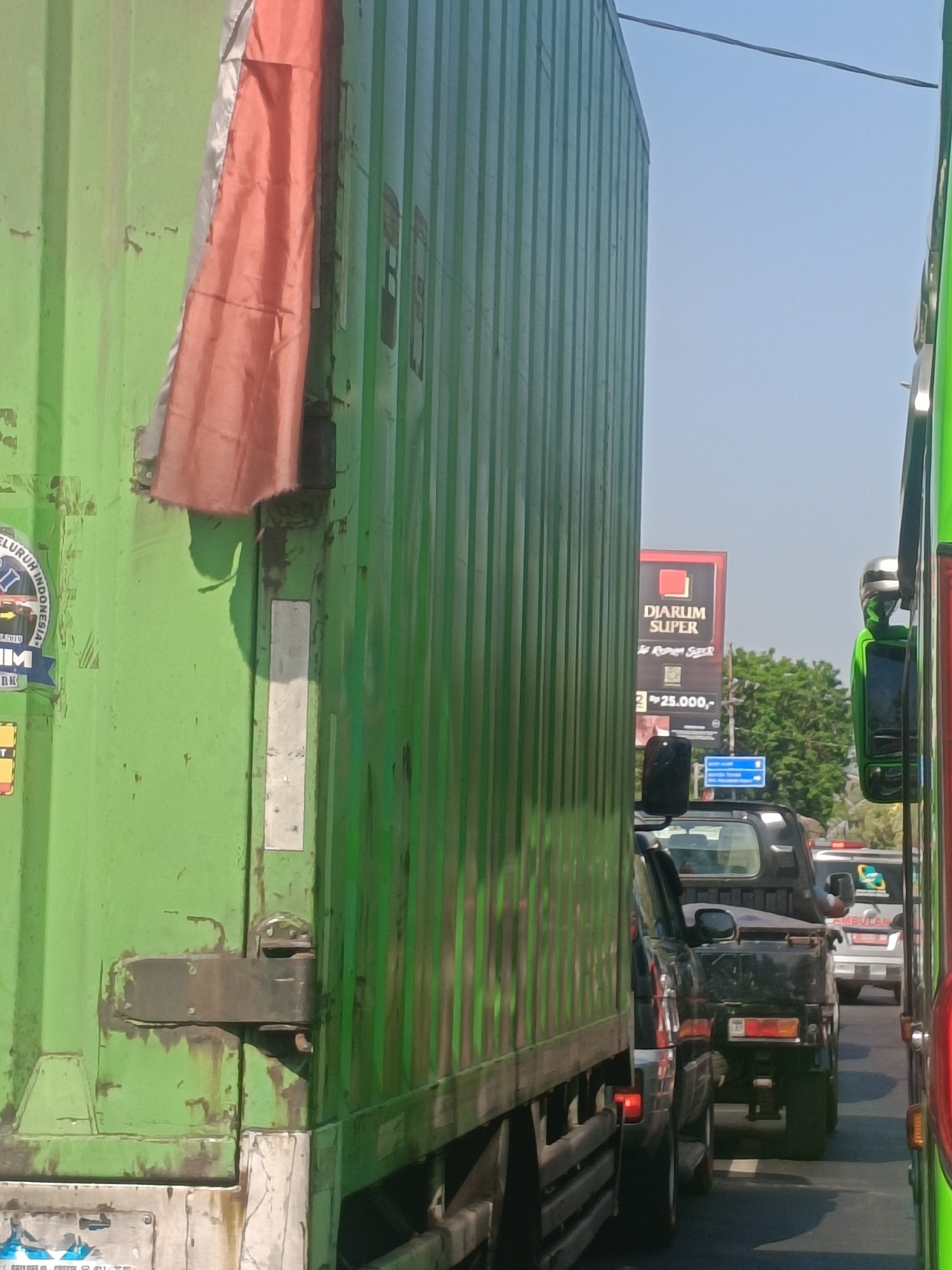The image shows a traffic jam on a sunny day in Kembangan, Indonesia. The foreground is dominated by the side of a large green container truck, with an orange tarp hanging from its upper section. To the right, a bright green bus is partially visible. In the middle ground, several vehicles are stopped in traffic, including a black pickup truck. Behind the vehicles, a billboard advertising "DIARLUM SUPER" is visible, along with trees and a clear blue sky. Road signs are also present. The scene depicts a typical urban traffic congestion scenario.