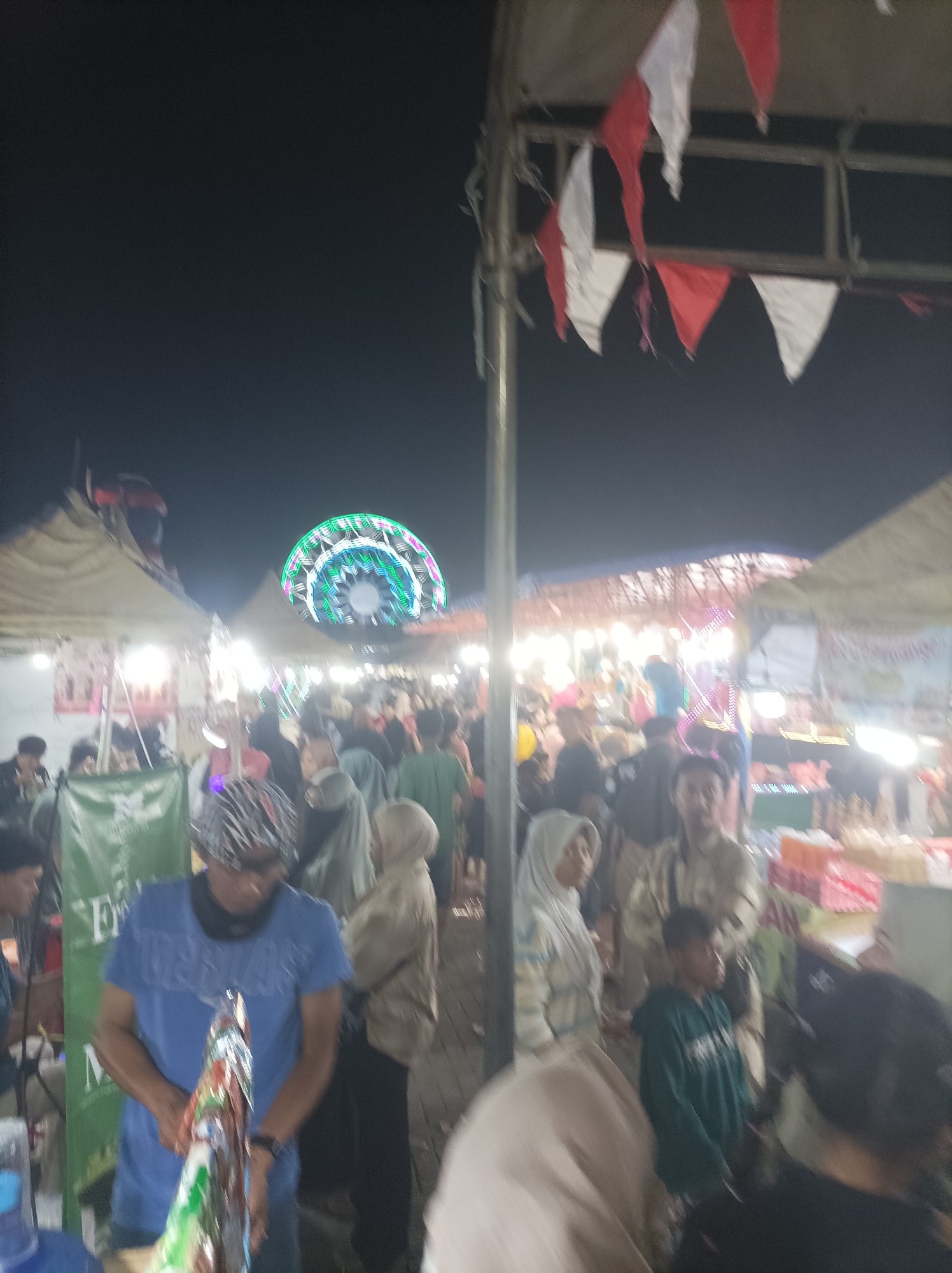 The image depicts a bustling night market scene in Cirebon, Indonesia. A Ferris wheel, illuminated with colorful lights, dominates the background. People are seen milling about, browsing stalls, and interacting with each other. Tents and temporary structures are set up, indicating a temporary market or festival. Red and white triangular flags are strung across some of the structures, adding to the festive atmosphere. The overall impression is one of a lively community gathering.