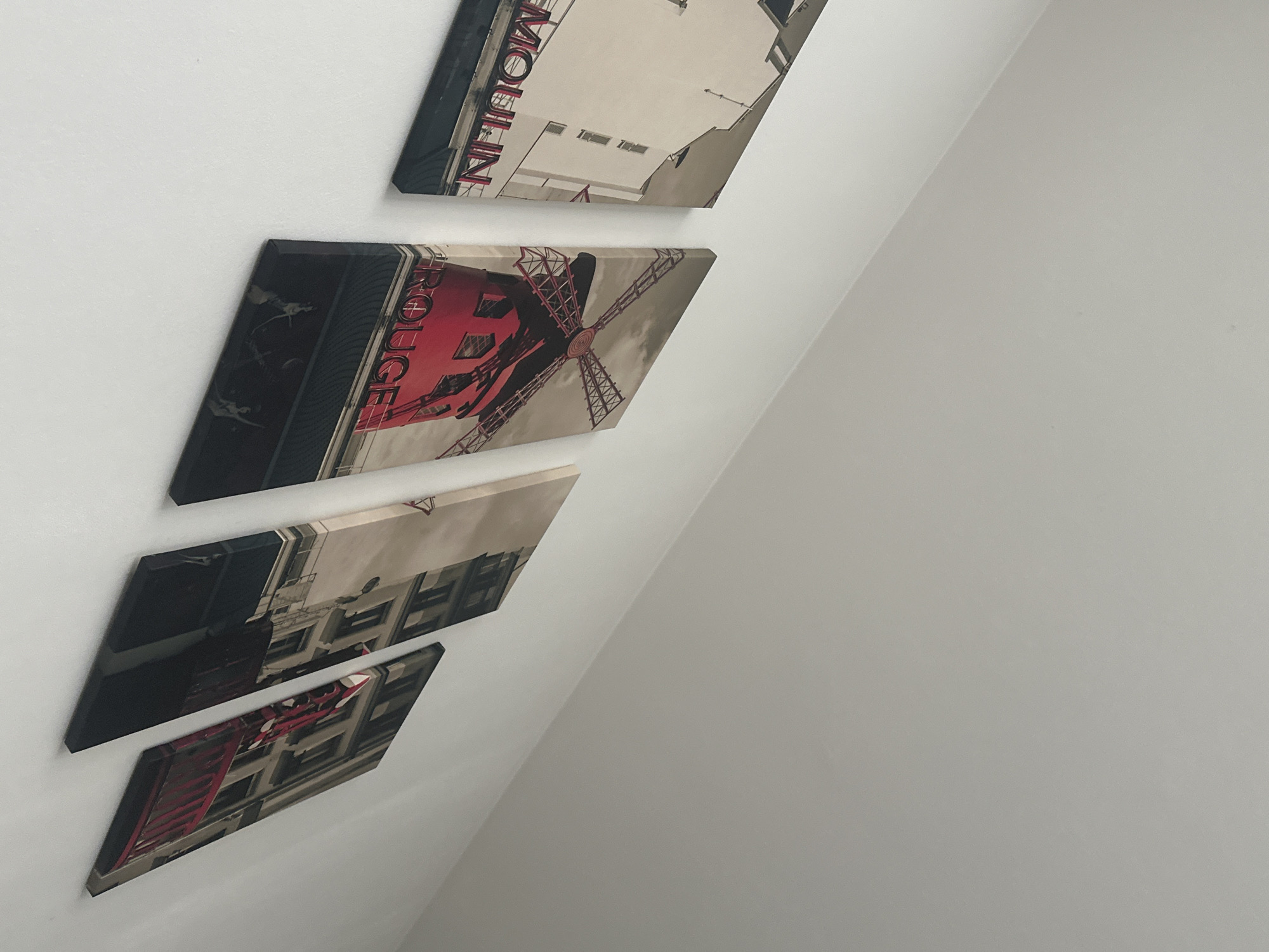 The image shows four canvas prints arranged diagonally on a white wall. The prints depict the Moulin Rouge in Paris. The top print shows the word "MOULIN" vertically. The second print shows the red windmill. The two bottom prints show the building's facade. The scene is indoors. There are no people present. The location is not Varna, Bulgaria, but Paris, France.