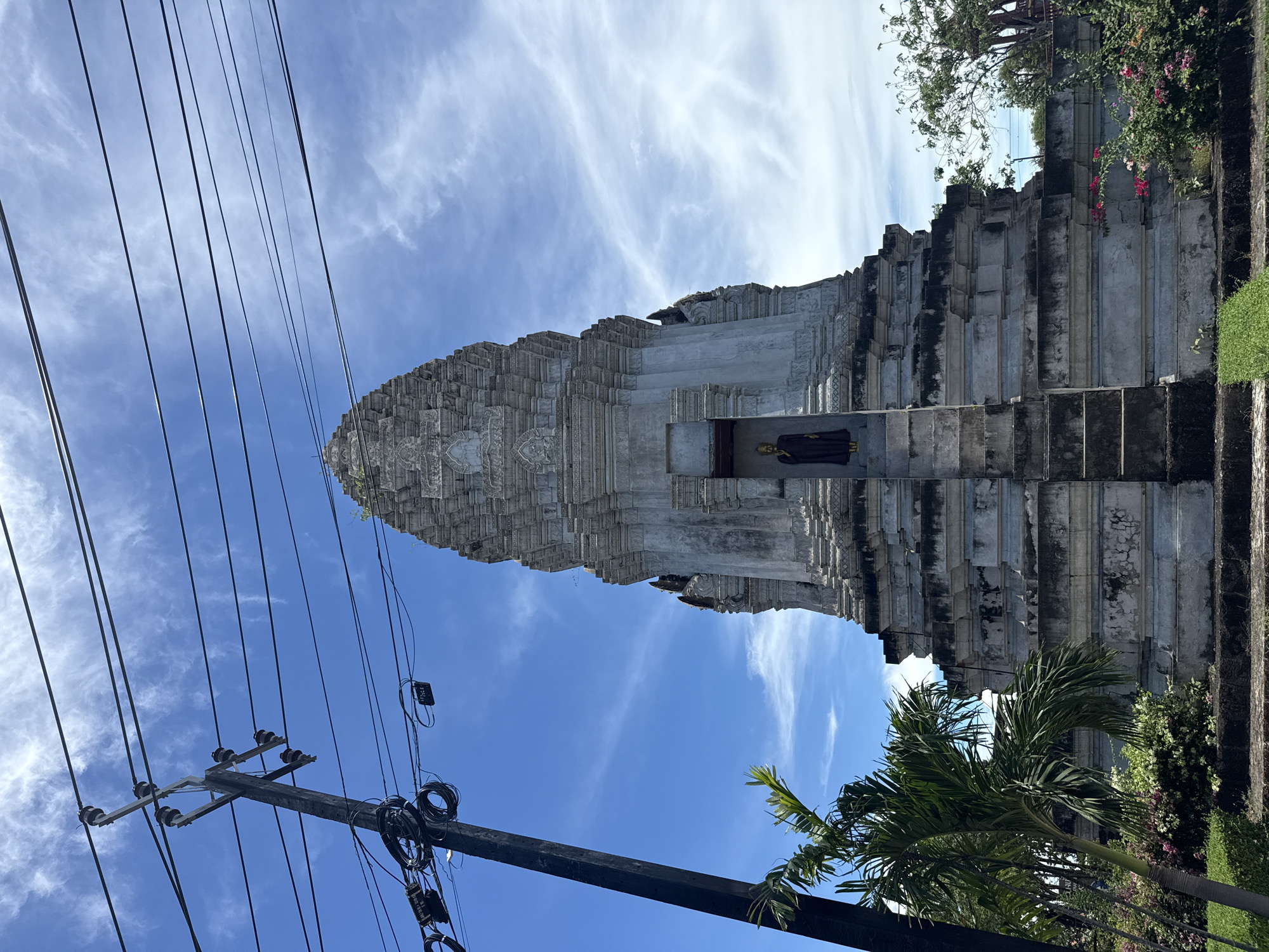 An upward, vertically-oriented shot captures a tall, gray stone structure akin to a stepped pyramid or tower under a blue sky with streaks of white clouds. A statue of a robed figure stands in a recess near the base. To the left, a utility pole and overhead wires extend into the sky, appearing to obscure parts of the structure. Green plants and palm trees are situated at the base of the building on the right side. This is located in Bang Pu Subdistrict Municipality, Thailand.