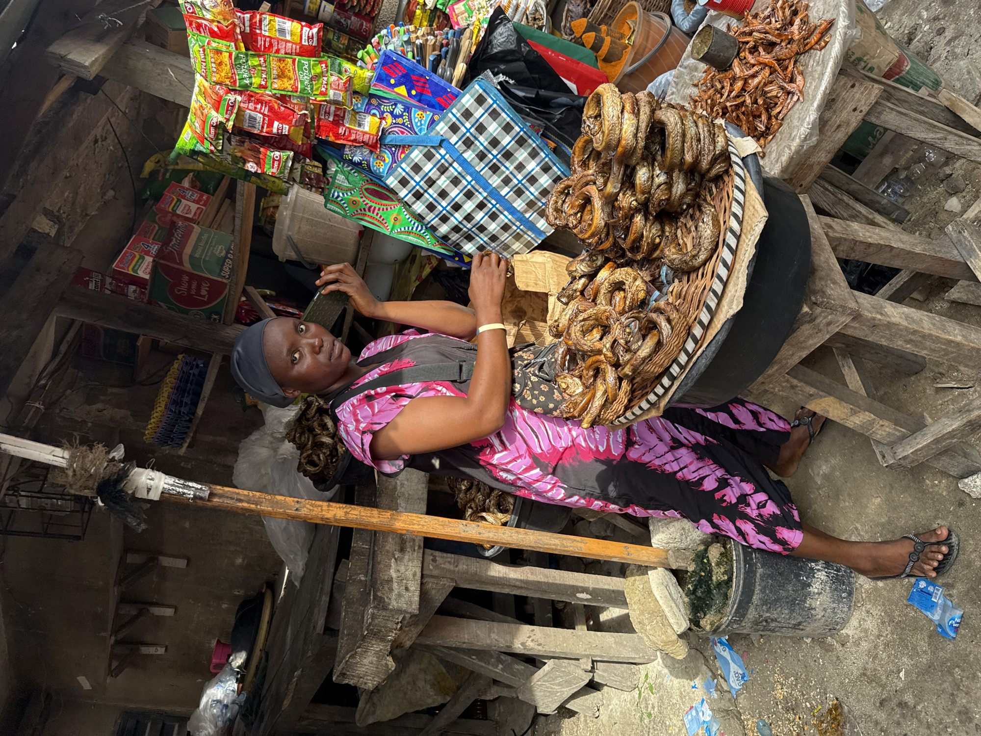 An image captures a scene from a market in Lekki, Nigeria, featuring a female vendor seated amidst her wares. The woman, wearing a patterned pink and black dress and a dark headscarf, looks directly at the viewer with a neutral expression. She is seated on the ground, with her right arm resting on a wooden structure supporting her stall.
Her stall displays a variety of goods. Prominently featured is a large woven basket filled with numerous rings of what appears to be smoked or dried fish, piled high. Next to this, on a separate platform, are quantities of dried small shrimp or crayfish. Behind these items, shelves and hangers hold various packaged products, including "MINI PACK" items, "Ikom Oil," "Instant Noodles," "SUPER DA," and products with the brand "Show SOME Love" and "POZ." Several colorful reusable shopping bags, including one with a blue and white plaid pattern, are also visible.
The market environment appears rustic, with rough wooden beams and structures forming the stall's framework. The ground is a mix of concrete and dirt, with some scattered debris, including plastic wrappers and a black bucket. Natural daylight illuminates the scene, suggesting it is daytime. The overall setting is characteristic of a bustling local market.