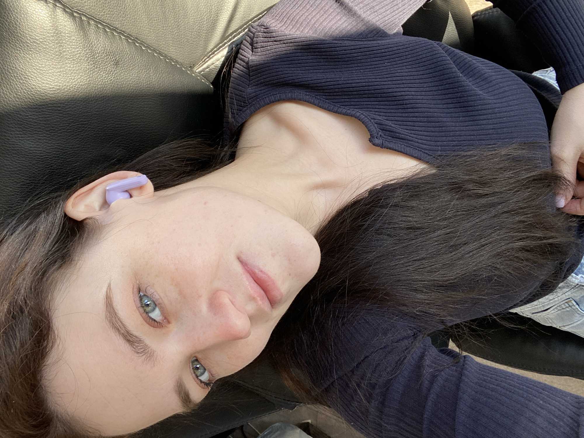 An overhead, slightly tilted shot captures a young person reclining in bright daylight, likely in Chaikovsky, Russia. The individual, appearing female, gazes directly at the camera with clear blue-grey eyes and a neutral to slightly pleasant expression. They have long, dark hair, some of which is draped across their chest and the dark surface they are lying on.
The subject is wearing a dark blue or black ribbed long-sleeved top, and glimpses of light-colored denim pants or shorts are visible on the right side of the frame. A lavender or light purple wireless earbud is visible in their right ear. They are resting on a dark, textured surface, possibly leather, which shows strong highlights and shadows from the sunlight, suggesting an outdoor or brightly lit indoor setting. The specific location cues are limited to this textured surface.
The activity appears to be posing for a selfie or a casual portrait while relaxing. The strong sunlight indicates it is daytime, under clear weather conditions. No visible text is present in the image.
